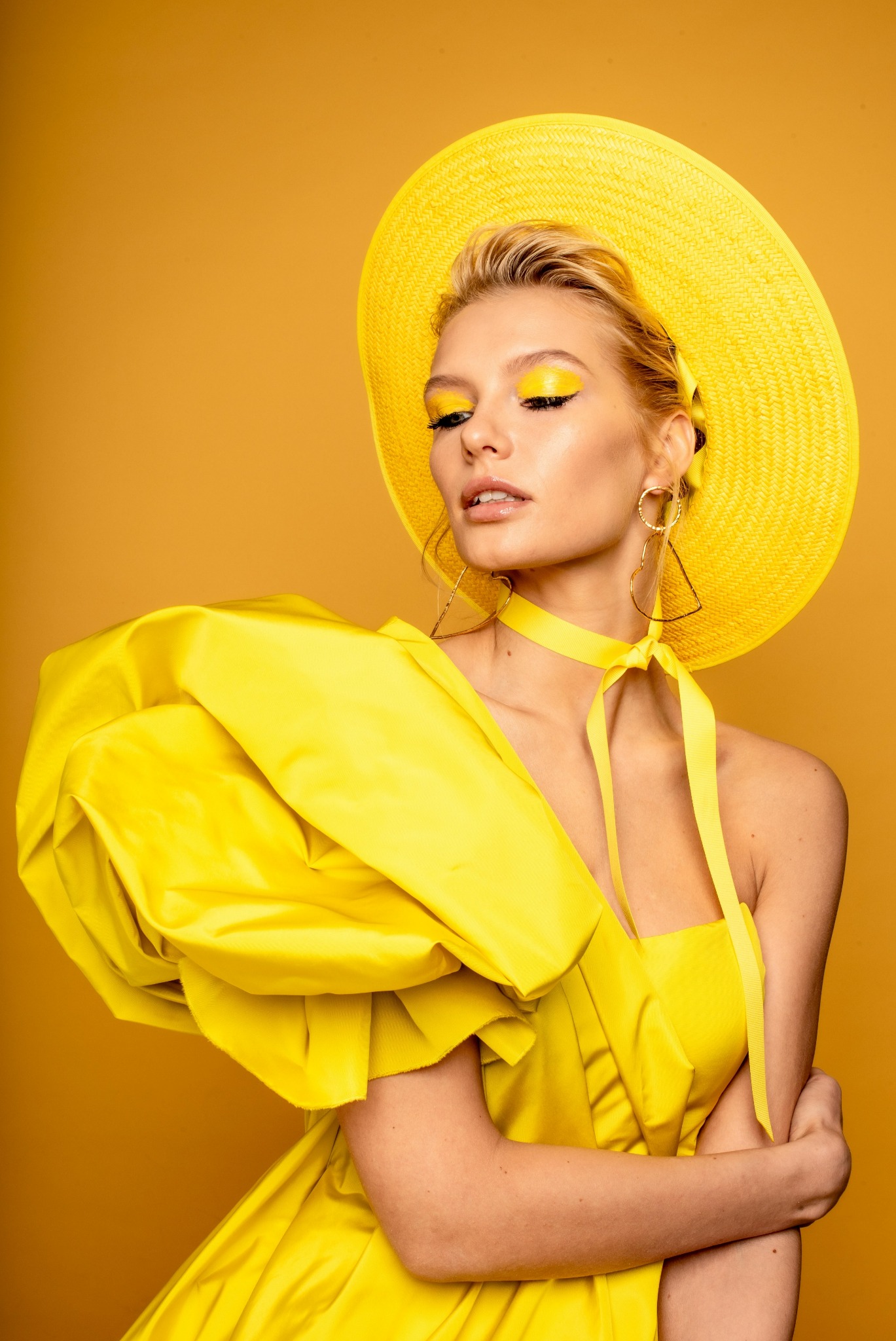 Portret kobiety w żółtym kapeluszu i sukni z bufkami, z żółtym makijażem oczu i kolczykami w kształcie serc, na jednolitym żółtym tle.