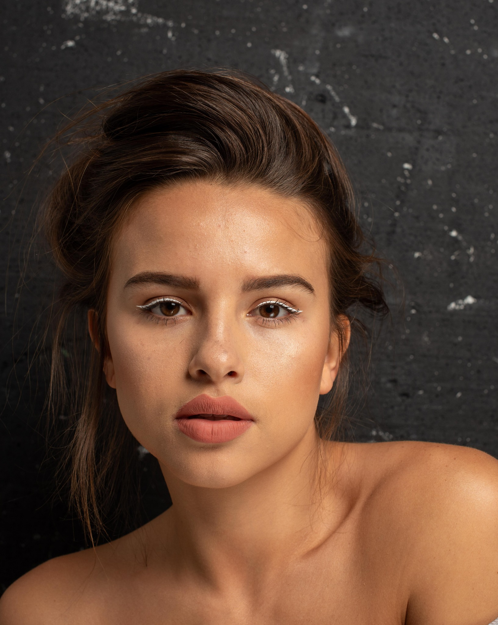 Portret młodej kobiety z delikatnym makijażem, białym eyelinerem i matową pomadką na tle ciemnej, teksturowanej ściany.