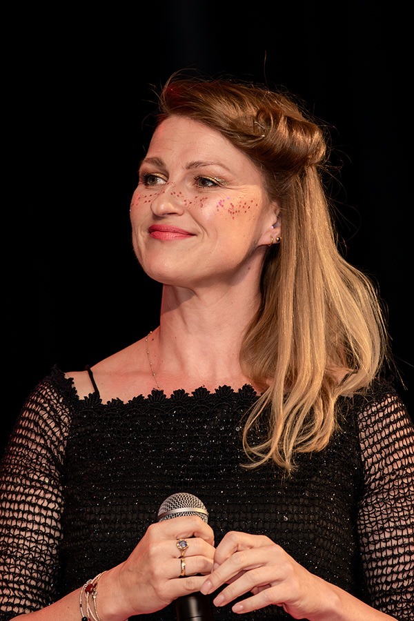 Uśmiechnięta kobieta z brokatowymi piegami, trzymająca mikrofon, w czarnej, błyszczącej sukience z koronkowymi rękawami, na czarnym tle.