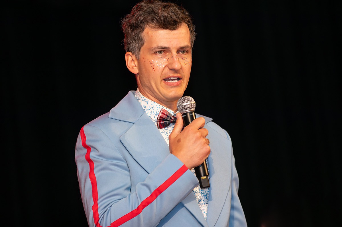 Mężczyzna w błękitnym garniturze z czerwoną lamówką, muszką w kratę i brokatem na twarzy trzyma mikrofon na czarnym tle.