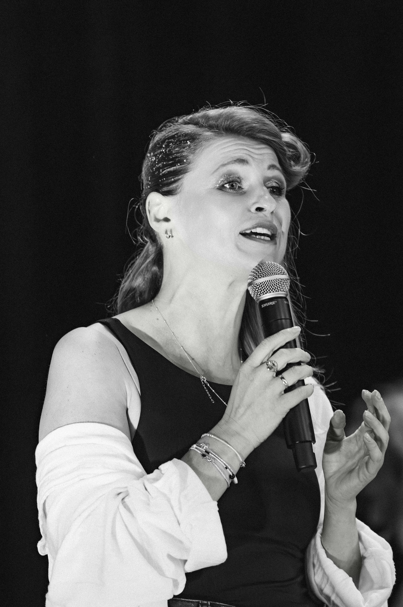 Kobieta w czarno-białym zdjęciu, trzymająca mikrofon Shure, z błyszczącymi drobinkami we włosach i licznymi bransoletkami na nadgarstku.