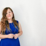 Portret uśmiechniętej kobiety w niebieskiej sukience na jasnym tle, z rękami splecionymi przed sobą, delikatny makijaż, długie, falowane, brązowe włosy opadające na ramiona.