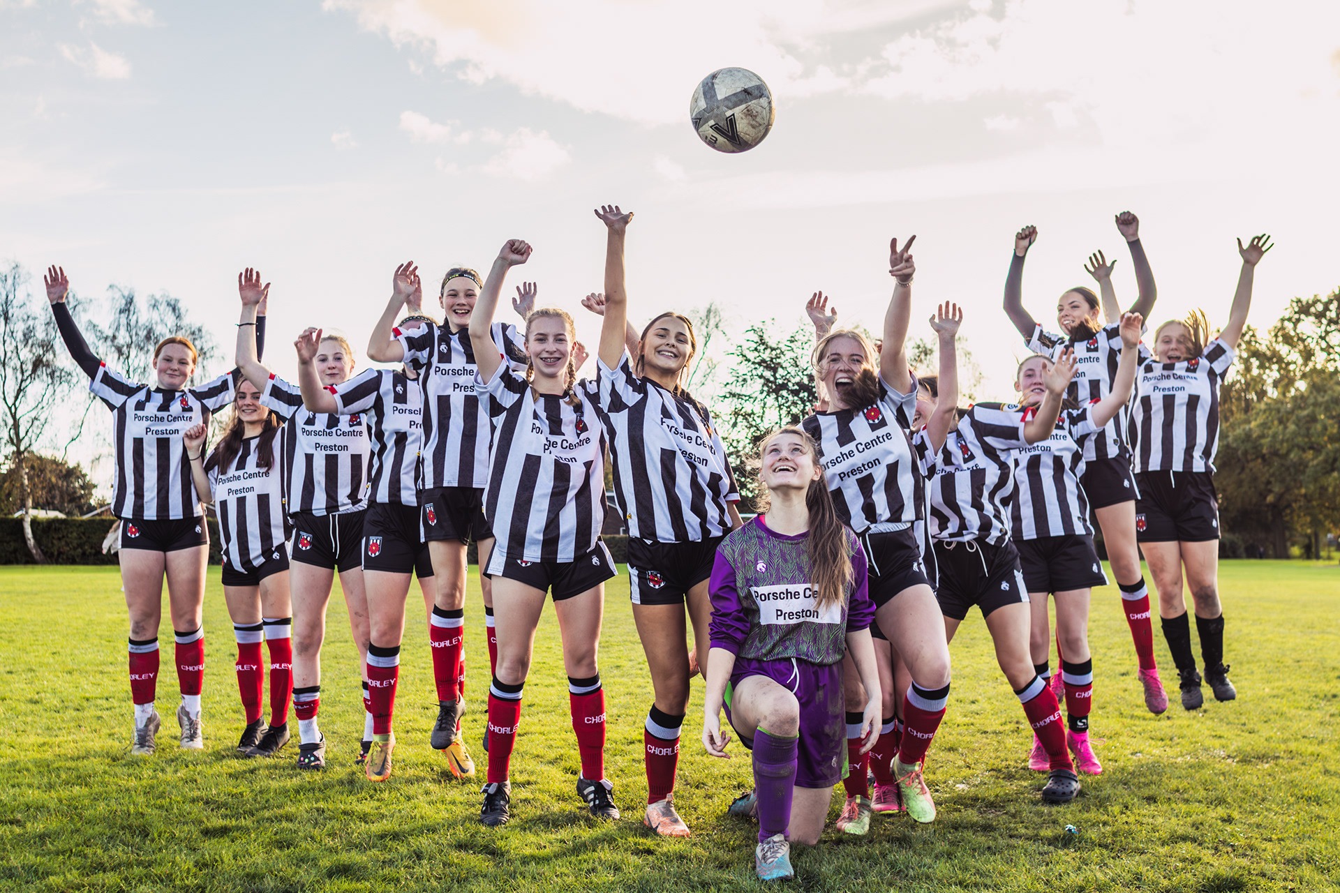 Drużyna piłkarska kobiet w czarno-białych strojach z logo sponsora, skacząca z radości na zielonej murawie, z piłką w powietrzu i jedną zawodniczką w fioletowym stroju klęczącą na pierwszym planie.