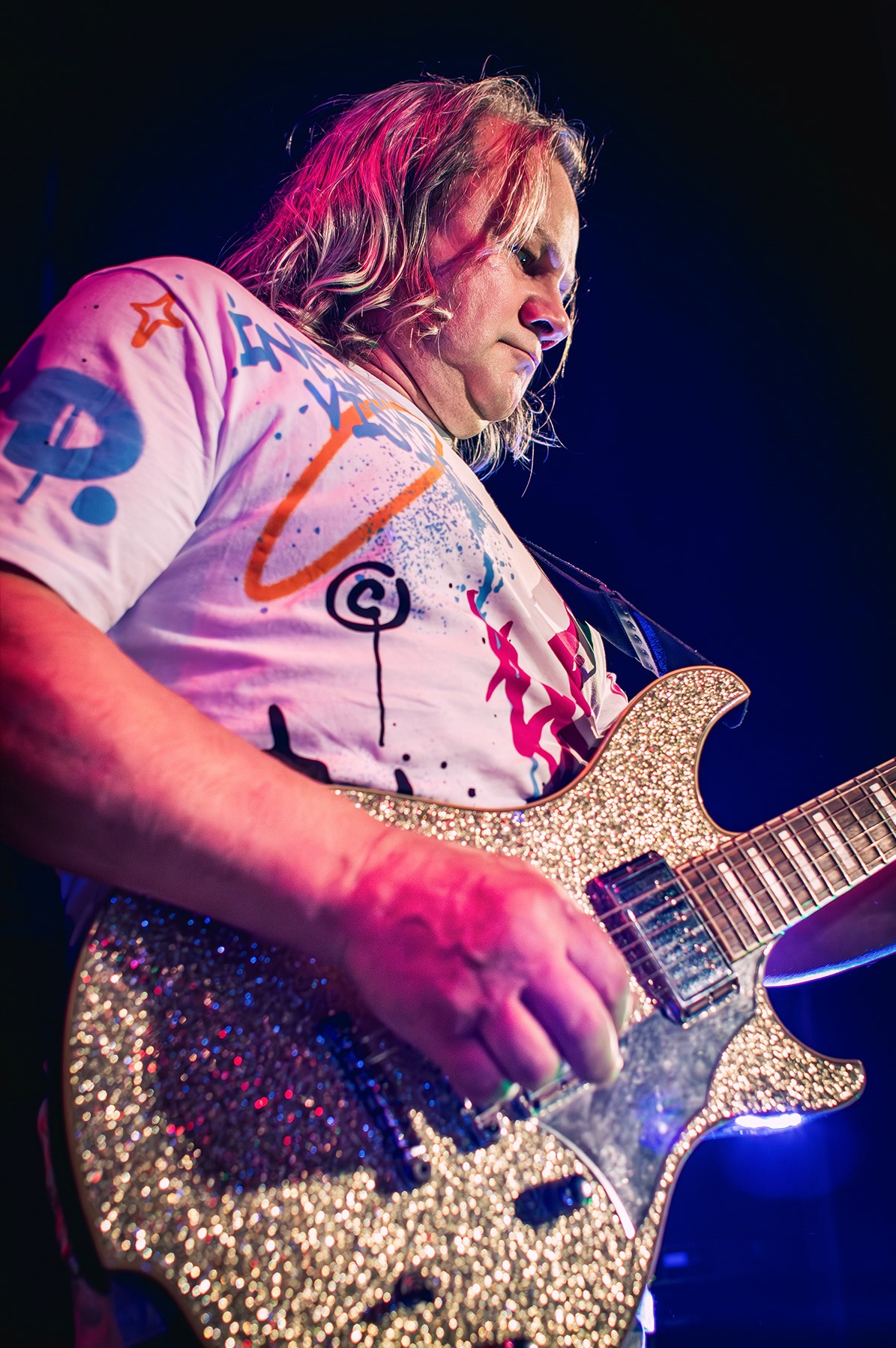 Muzyk z długimi blond włosami gra na błyszczącej, złotej gitarze elektrycznej w trakcie koncertu, ujęcie od dołu w mocnym oświetleniu scenicznym.