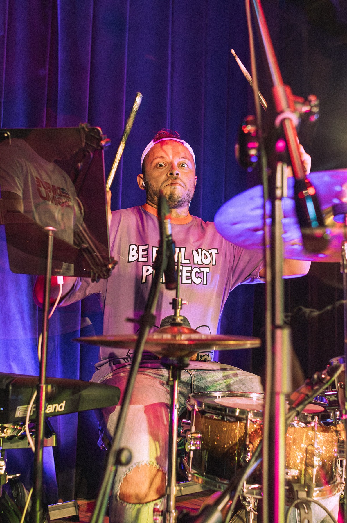 Perkusista z wyrazem zaskoczenia na twarzy, w koszulce z napisem 'Be Real Not Perfect', w trakcie występu na scenie z błękitnymi zasłonami w tle.