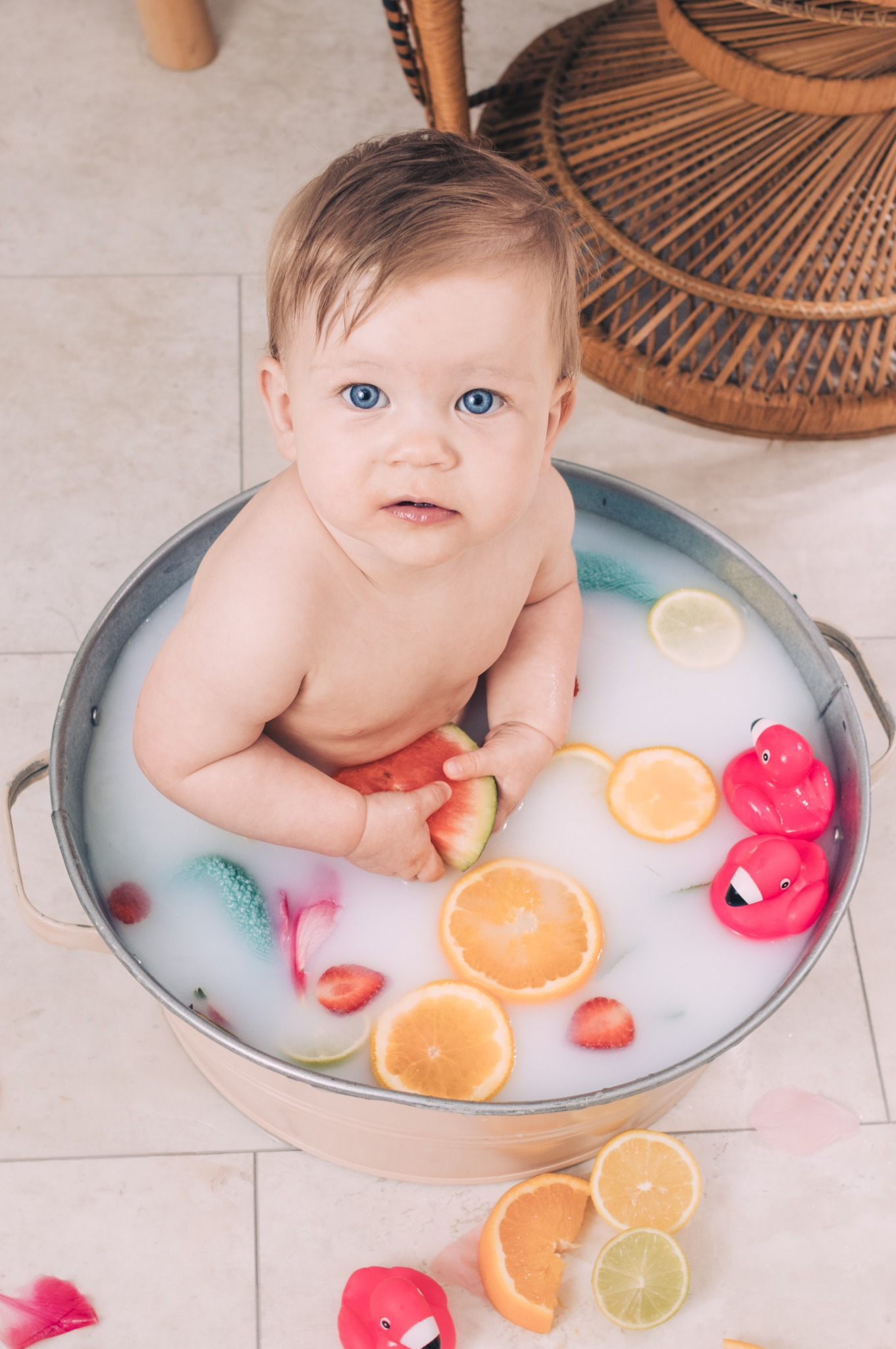 Niemowlę z niebieskimi oczami siedzi w metalowej wanience wypełnionej mleczną wodą z pływającymi plasterkami pomarańczy, cytryny, truskawek, arbuza, gumowymi flamingami i dekoracyjnymi płatkami...