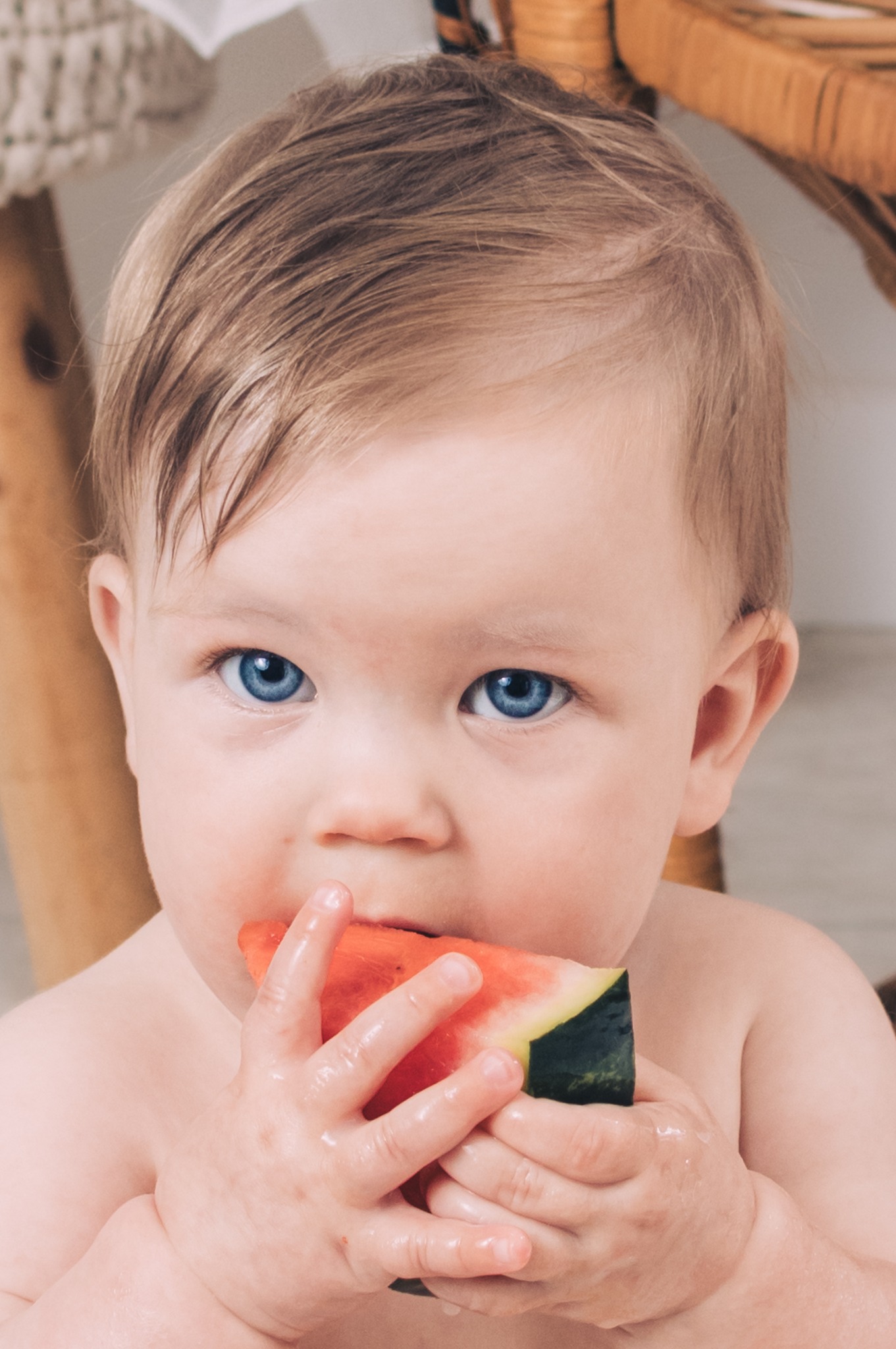 Niemowlę z jasnymi włosami i niebieskimi oczami, trzymające w dłoniach duży kawałek arbuza i gryzące go, z widocznymi kroplami soku na dłoniach, na rozmytym tle jasnego wnętrza.