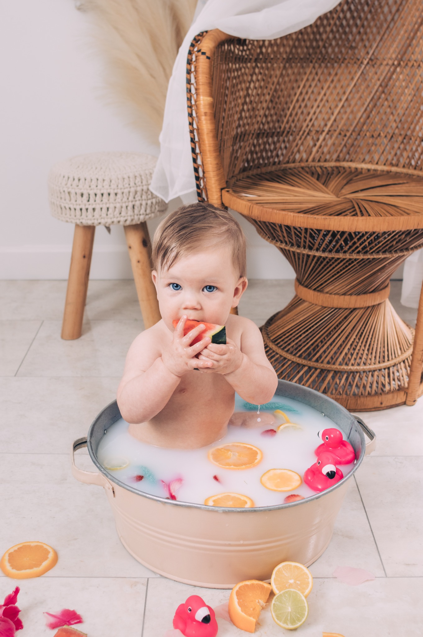 Niemowlę z niebieskimi oczami siedzi w metalowej wanience wypełnionej mleczną wodą z plasterkami pomarańczy i gumowymi flamingami, zajadając arbuza na tle wiklinowego krzesła i taboretu.