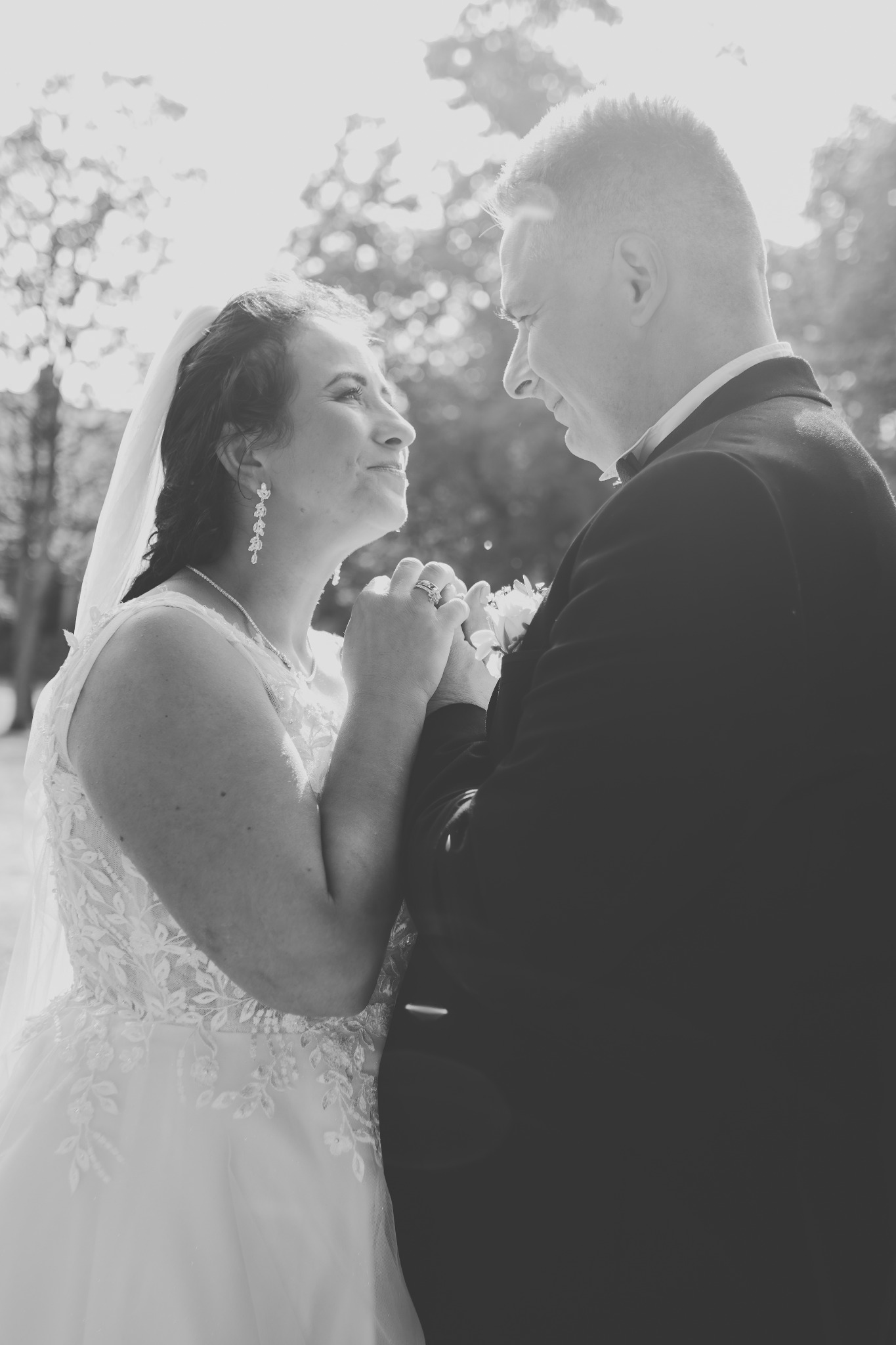 Czarno-białe zdjęcie pary młodej trzymającej się za ręce, panna młoda w koronkowej sukni i z welonem spogląda na pana młodego w smokingu, w tle rozmyte drzewa.