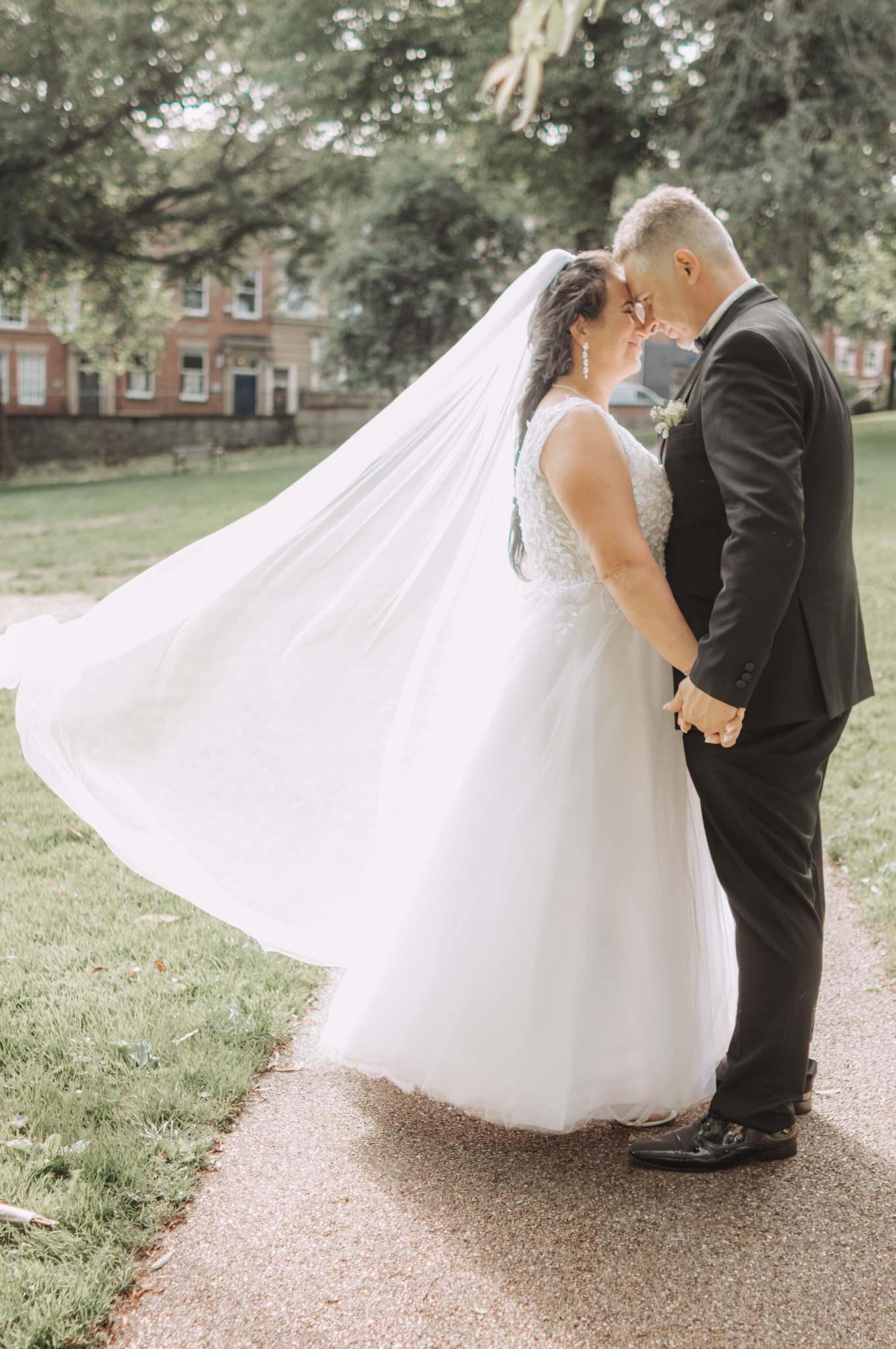 Para młoda, panna młoda w białej sukni i długim welonie, pan młody w czarnym garniturze, stoją na ścieżce w parku, dotykając się czołami.