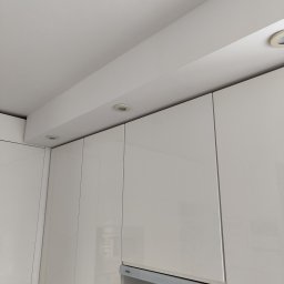 Przemysław Paziuk Front Home - Białe, lakierowane szafki kuchenne pod sufitem z zamontowanym oświetleniem punktowym, widoczny okap kuchenny marki Amica i panel ścienny z motywem górskim.