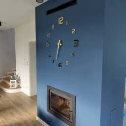 Przemysław Paziuk Front Home - Niebieska ściana z kominkiem, złotym zegarem naklejonym bezpośrednio na ścianę i kratką wentylacyjną u góry, widok na drewniane schody i lampion dekoracyjny w tle.