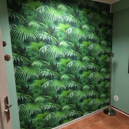 Przemysław Paziuk Front Home - Ściana pokryta tapetą z motywem tropikalnych liści, sąsiadująca z gładką, zieloną ścianą i brązową podłogą z terakoty. W rogu widoczna lampa podłogowa ze srebrną podstawą.