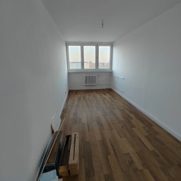 Przemysław Paziuk Front Home - Pusty pokój po remoncie z drewnianą podłogą, białymi ścianami i oknem z widokiem na miasto; na podłodze przy ścianie leżą panele podłogowe w opakowaniach.