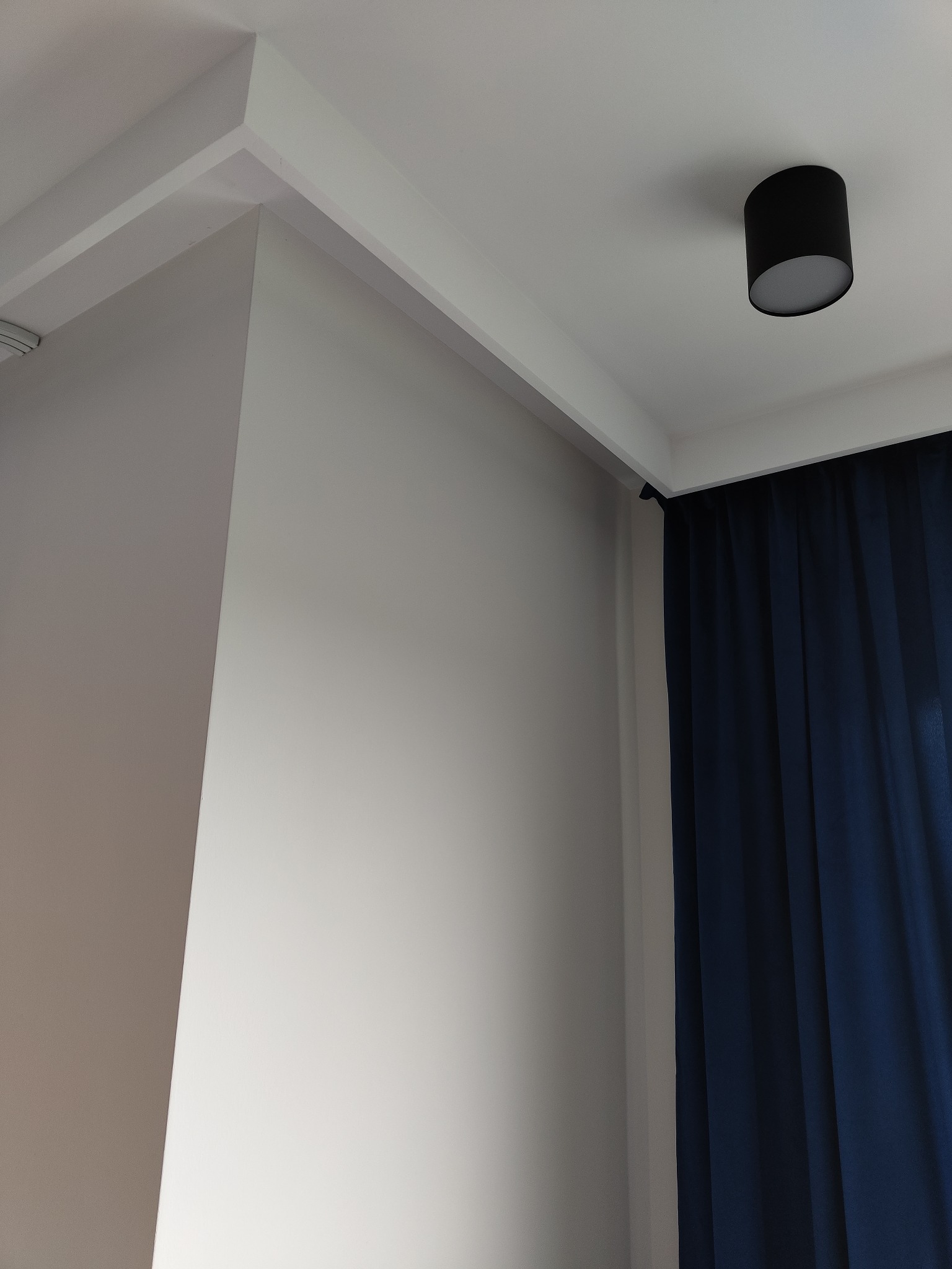 Pomalowany narożnik pokoju z eleganckim sufitem podwieszanym, oświetleniem punktowym i granatowymi zasłonami.