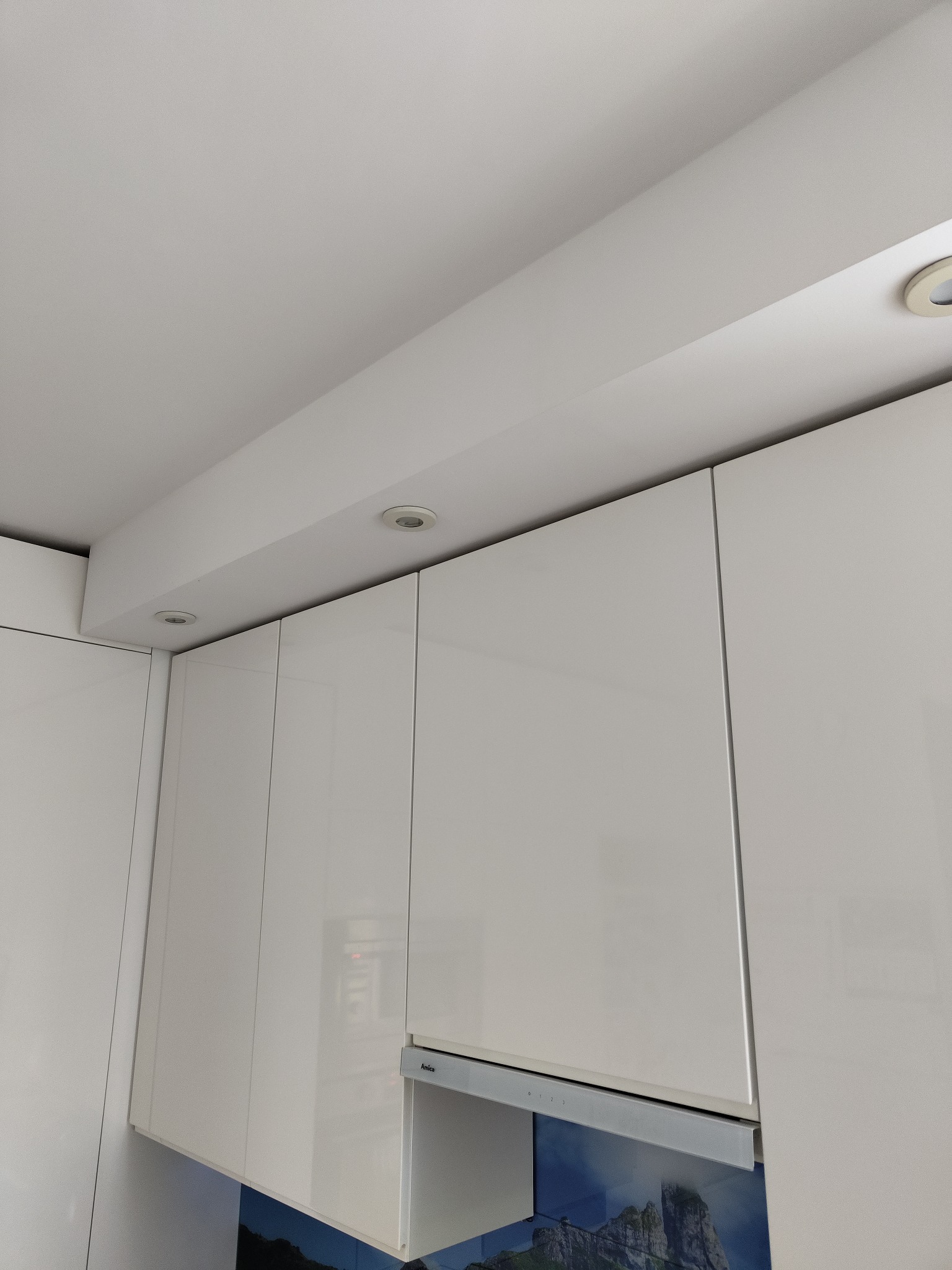 Białe, lakierowane szafki kuchenne pod sufitem z zamontowanym oświetleniem punktowym, widoczny okap kuchenny marki Amica i panel ścienny z motywem górskim.