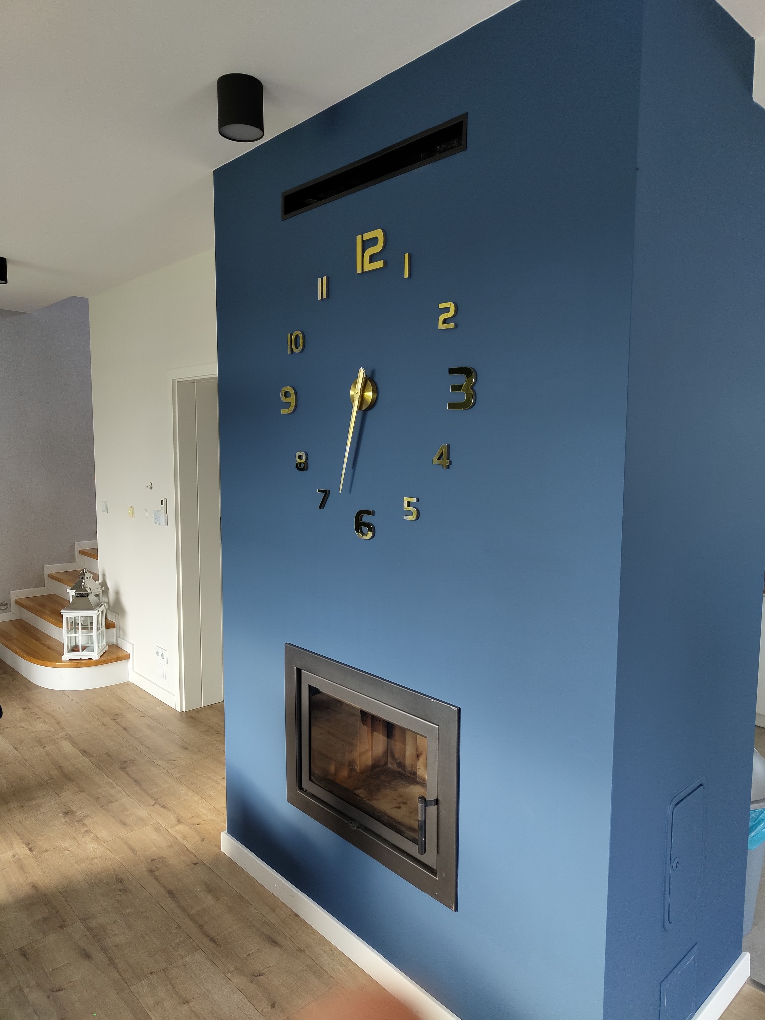 Niebieska ściana z kominkiem, złotym zegarem naklejonym bezpośrednio na ścianę i kratką wentylacyjną u góry, widok na drewniane schody i lampion dekoracyjny w tle.