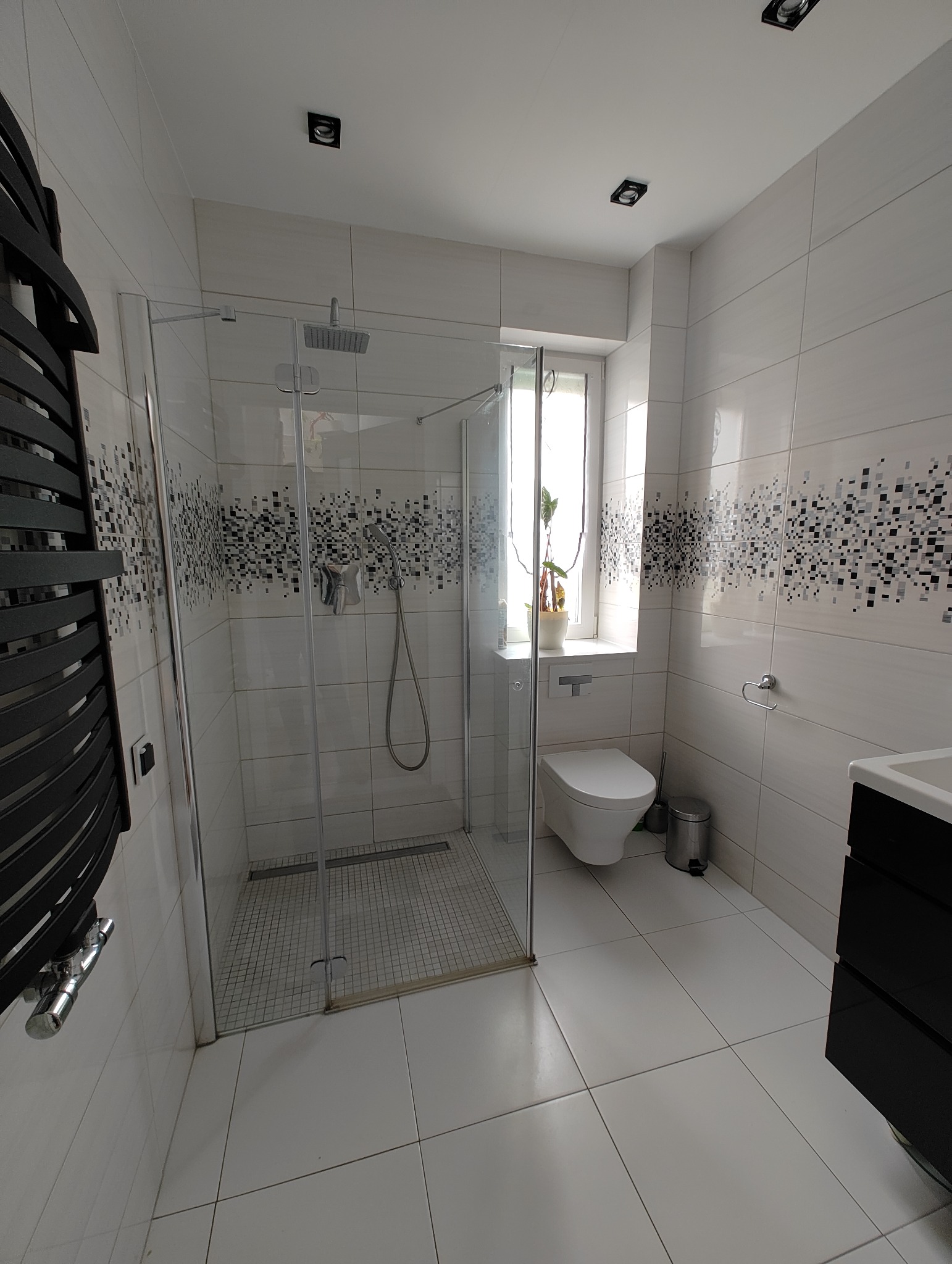 Nowoczesna łazienka z kabiną prysznicową typu walk-in, białymi płytkami z dekoracyjnym pasem mozaiki, podwieszaną toaletą i czarnym grzejnikiem drabinkowym.