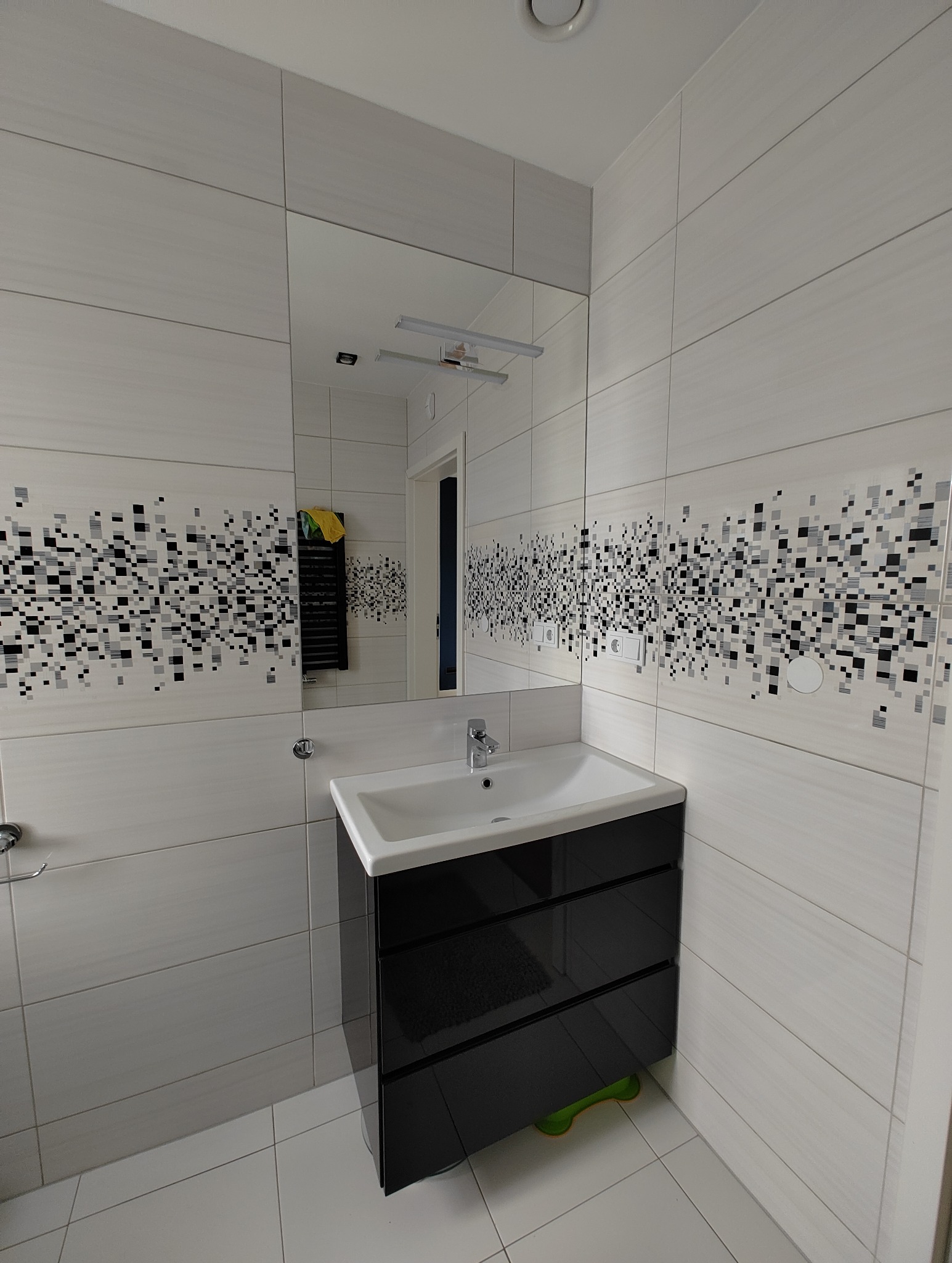 Nowoczesna łazienka z białymi płytkami w odcieniach szarości z motywem pikseli, czarna szafka pod umywalką z trzema szufladami, widoczne akcesoria łazienkowe i fragment grzejnika drabinkowego...