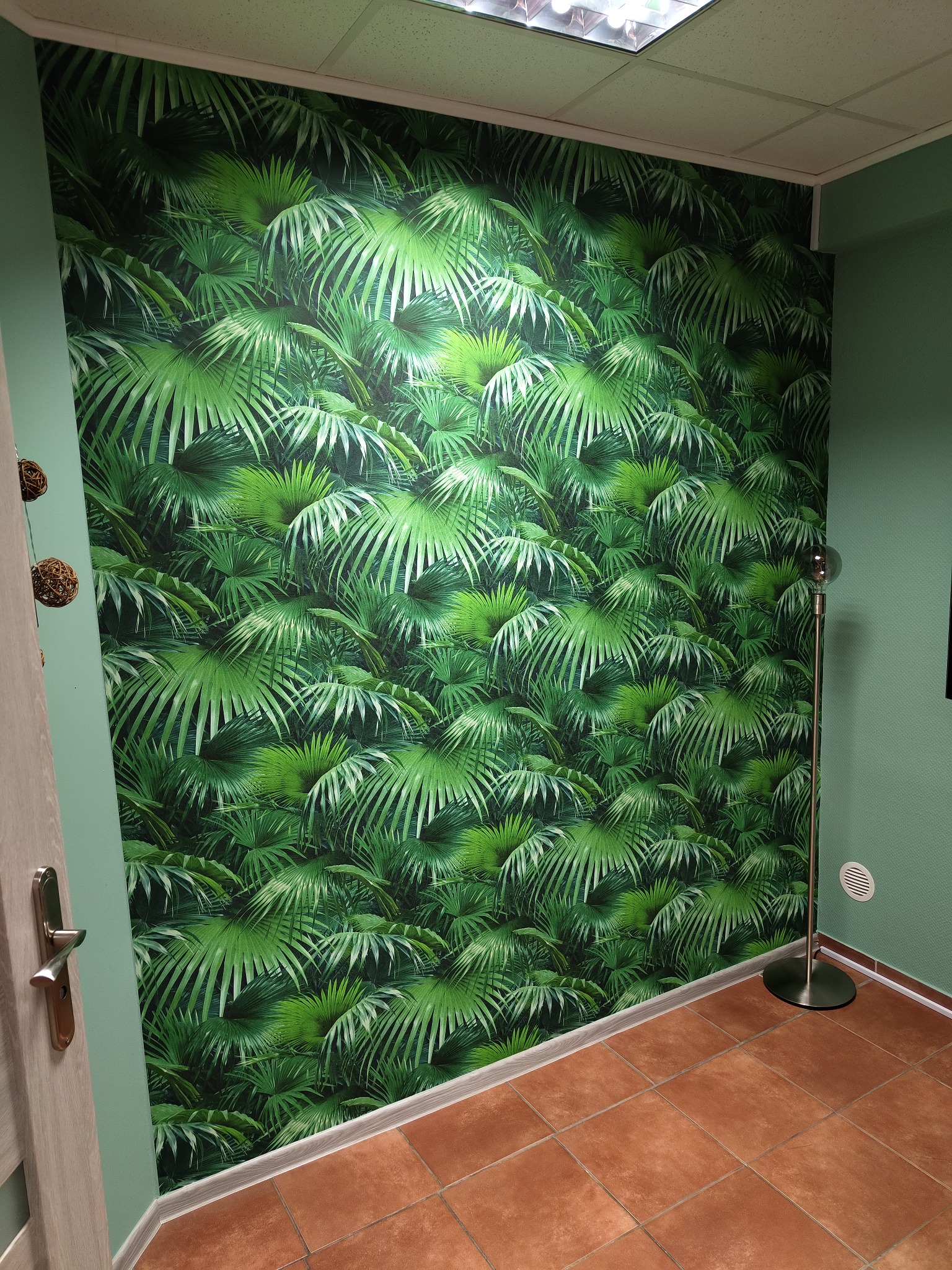 Ściana pokryta tapetą z motywem tropikalnych liści, sąsiadująca z gładką, zieloną ścianą i brązową podłogą z terakoty. W rogu widoczna lampa podłogowa ze srebrną podstawą.