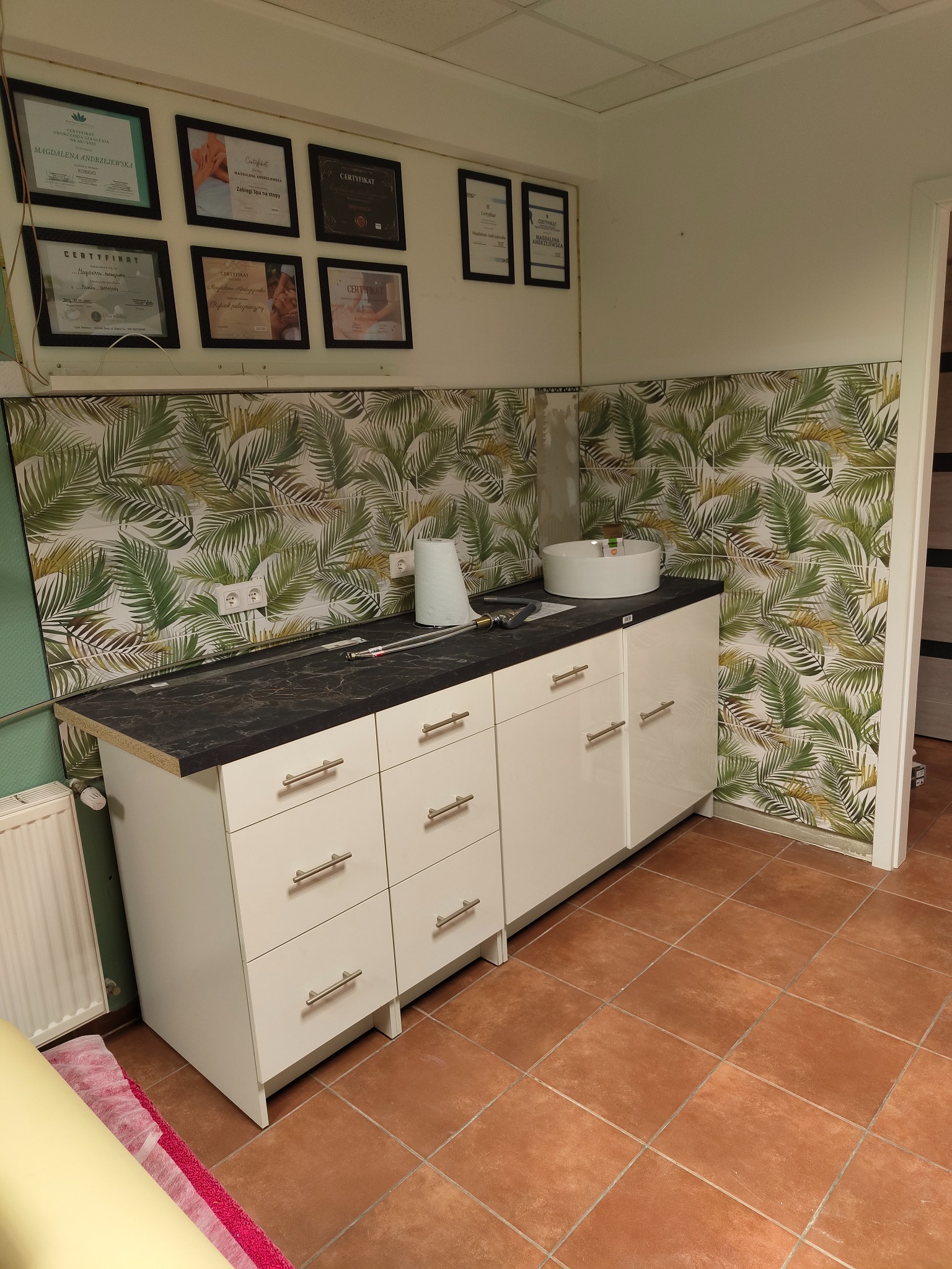 Łazienka w trakcie wykańczania z białymi szafkami pod umywalką, czarnym blatem, brązowymi płytkami podłogowymi i tapetą w tropikalne liście na ścianie, widoczne narzędzia i certyfikaty na ścianie.