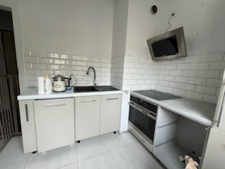 Kuchnia w trakcie wykańczania: beżowe szafki dolne z czarnym blatem i zlewozmywakiem, białe płytki ścienne typu metro z ciemną fugą, wiszący telewizor, niedokończona zabudowa piekarnika.