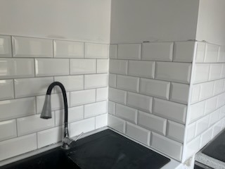 Białe, błyszczące płytki ułożone w cegiełkę na ścianie nad blatem kuchennym, widoczny fragment czarnego blatu i bateria umywalkowa z giętką wylewką.