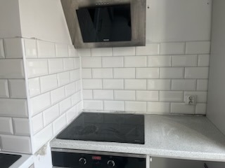 Białe, prostokątne płytki ułożone poziomo na ścianie nad blatem kuchennym, widoczny fragment płyty indukcyjnej i okap kuchenny ze stali nierdzewnej. Szara fuga. W tle gniazdko elektryczne.