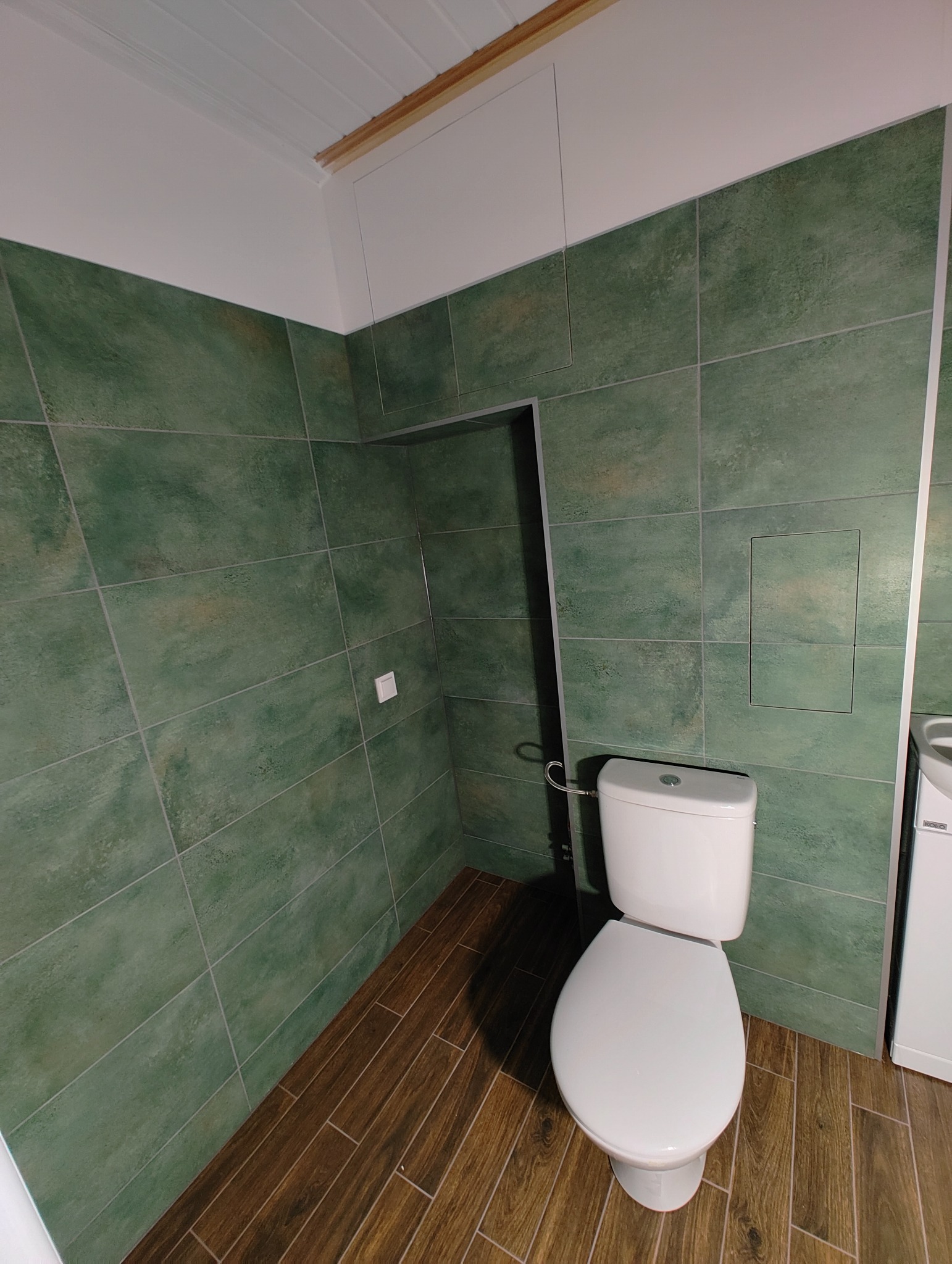 Wykończona łazienka z toaletą, zielonymi płytkami na ścianach i drewnopodobną podłogą. Widoczna klapka rewizyjna na ścianie i biały sufit z drewnianymi listwami.