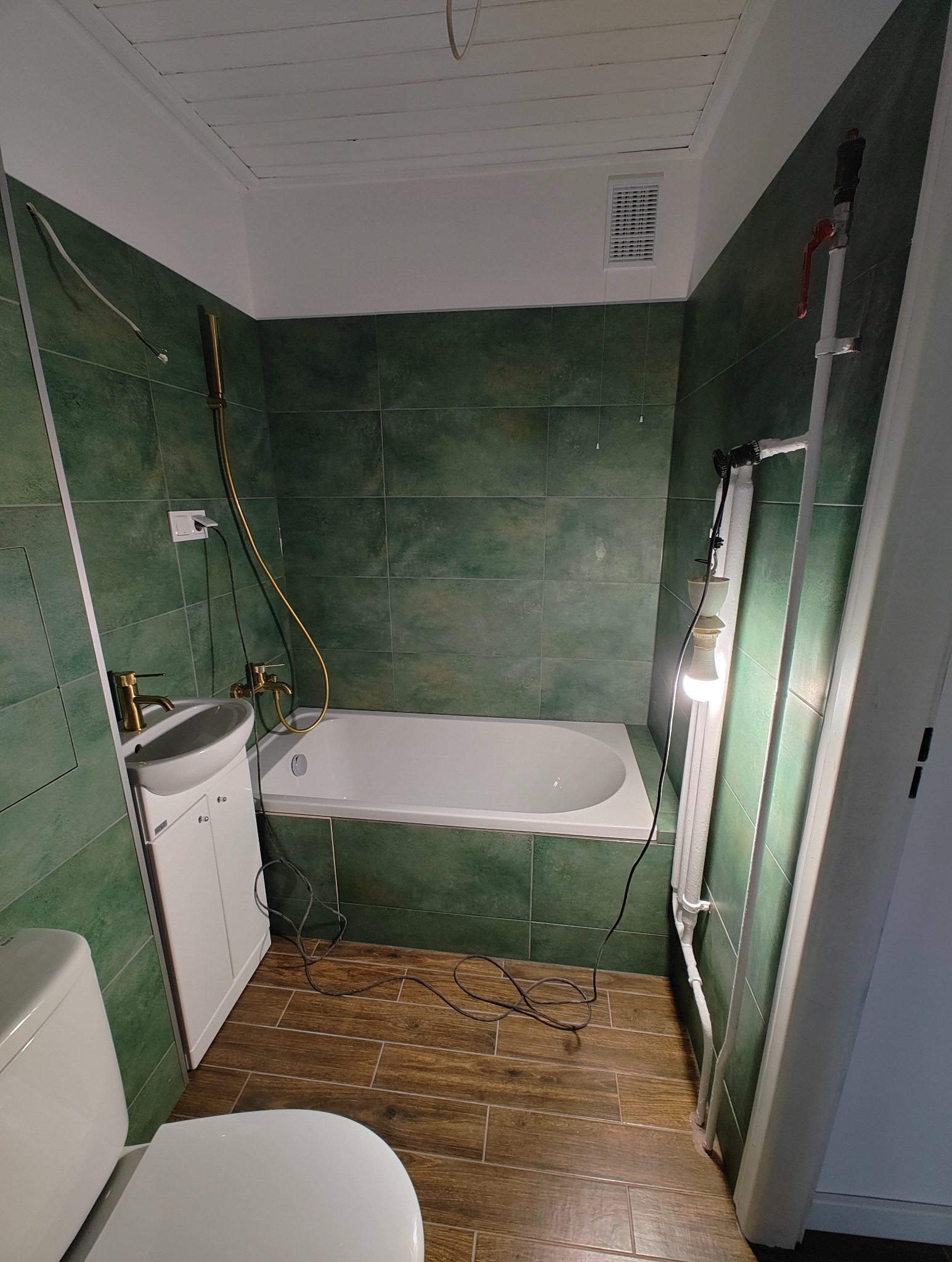 Wykończona łazienka z zielonymi płytkami, drewnopodobną podłogą, białą wanną, umywalką z szafką i toaletą; widoczne elementy instalacji, takie jak rury i grzejnik, oraz prowizoryczne oświetlenie...