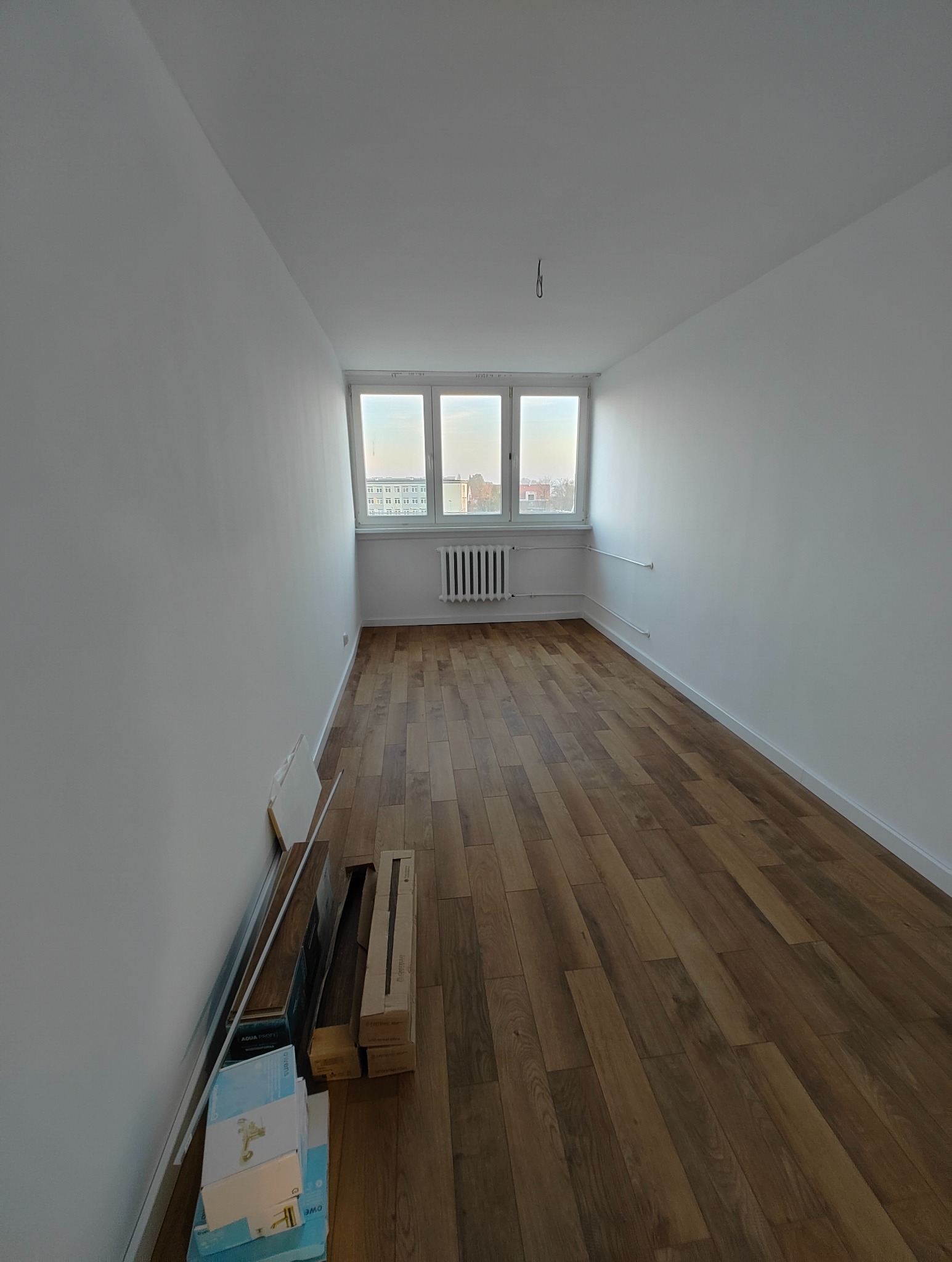 Pusty pokój po remoncie z drewnianą podłogą, białymi ścianami i oknem z widokiem na miasto; na podłodze przy ścianie leżą panele podłogowe w opakowaniach.