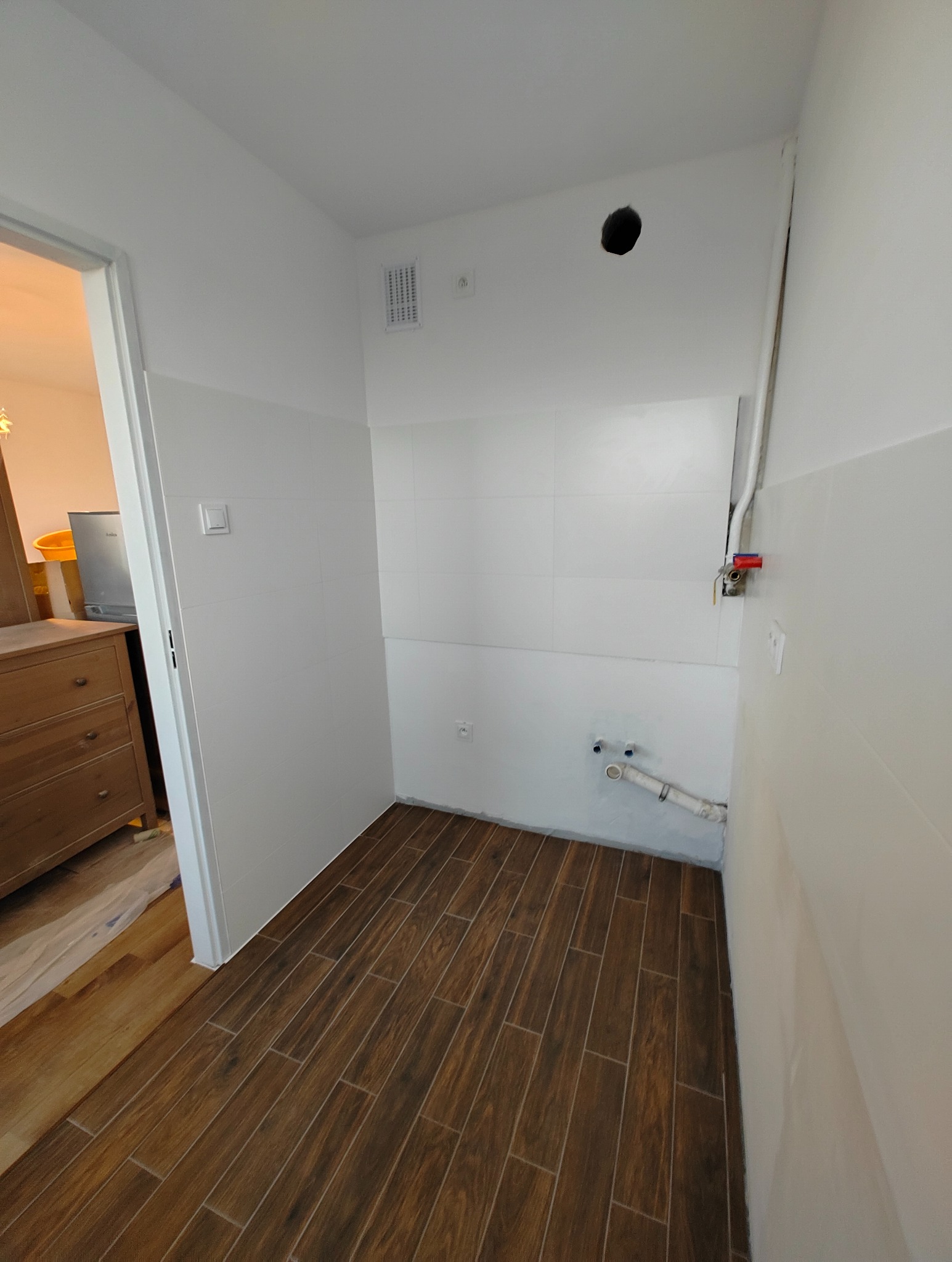 Wnętrze łazienki w trakcie remontu: białe płytki ścienne, brązowa podłoga imitująca drewno, widoczne rury hydrauliczne i otwór w suficie.