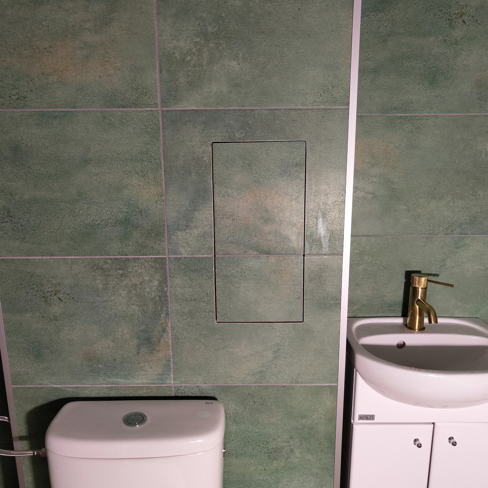 Łazienka po remoncie: zielone płytki z ukrytą klapą rewizyjną, biała toaleta i umywalka z baterią w kolorze złotym.