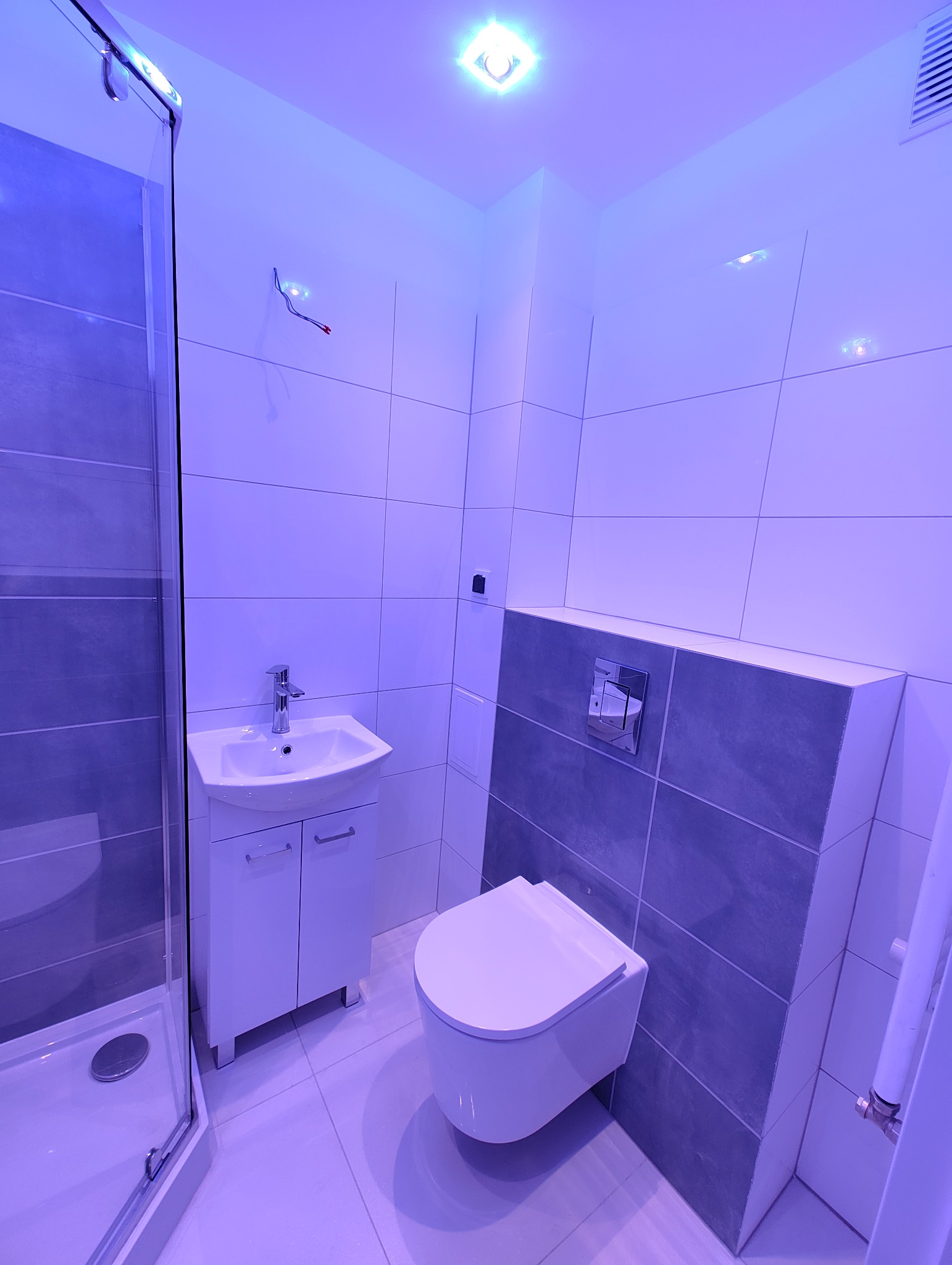 Nowoczesna łazienka z kabiną prysznicową, umywalką z szafką i podwieszanym WC, wykończona białymi i szarymi płytkami, oświetlona niebieskim światłem.