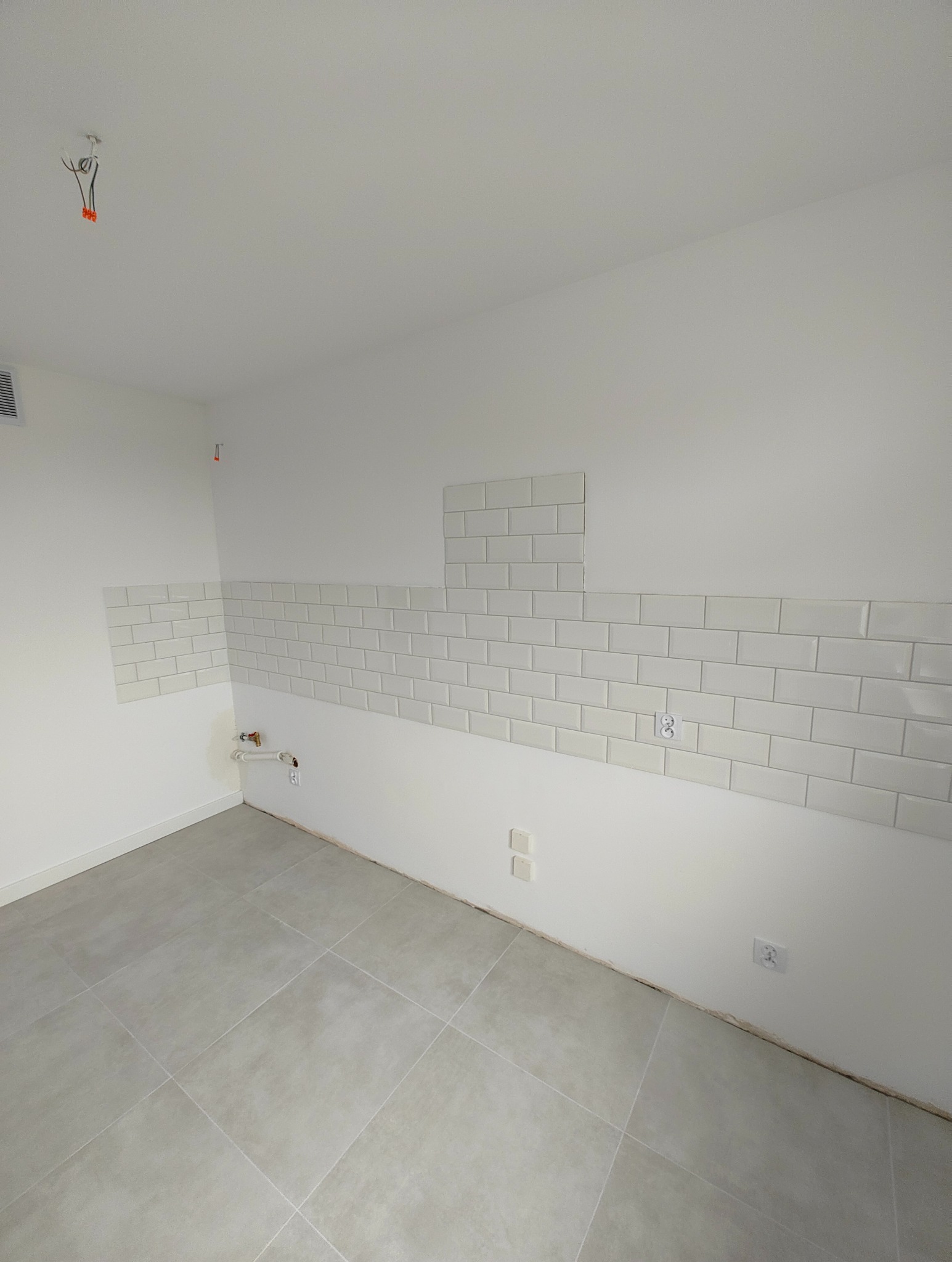 Wnętrze pomieszczenia w trakcie wykańczania: częściowo położone białe kafelki typu metro na ścianach, szara podłoga z płytek, widoczne puszki elektryczne i fragment instalacji wodnej.