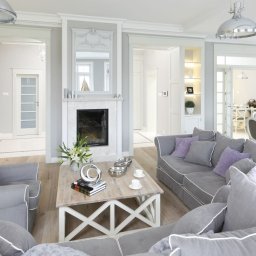 MBLux - Jasne, przestronne wnętrze salonu z kominkiem, szarymi sofami z fioletowymi poduszkami, drewnianym stolikiem kawowym i widokiem na jadalnię w tle.