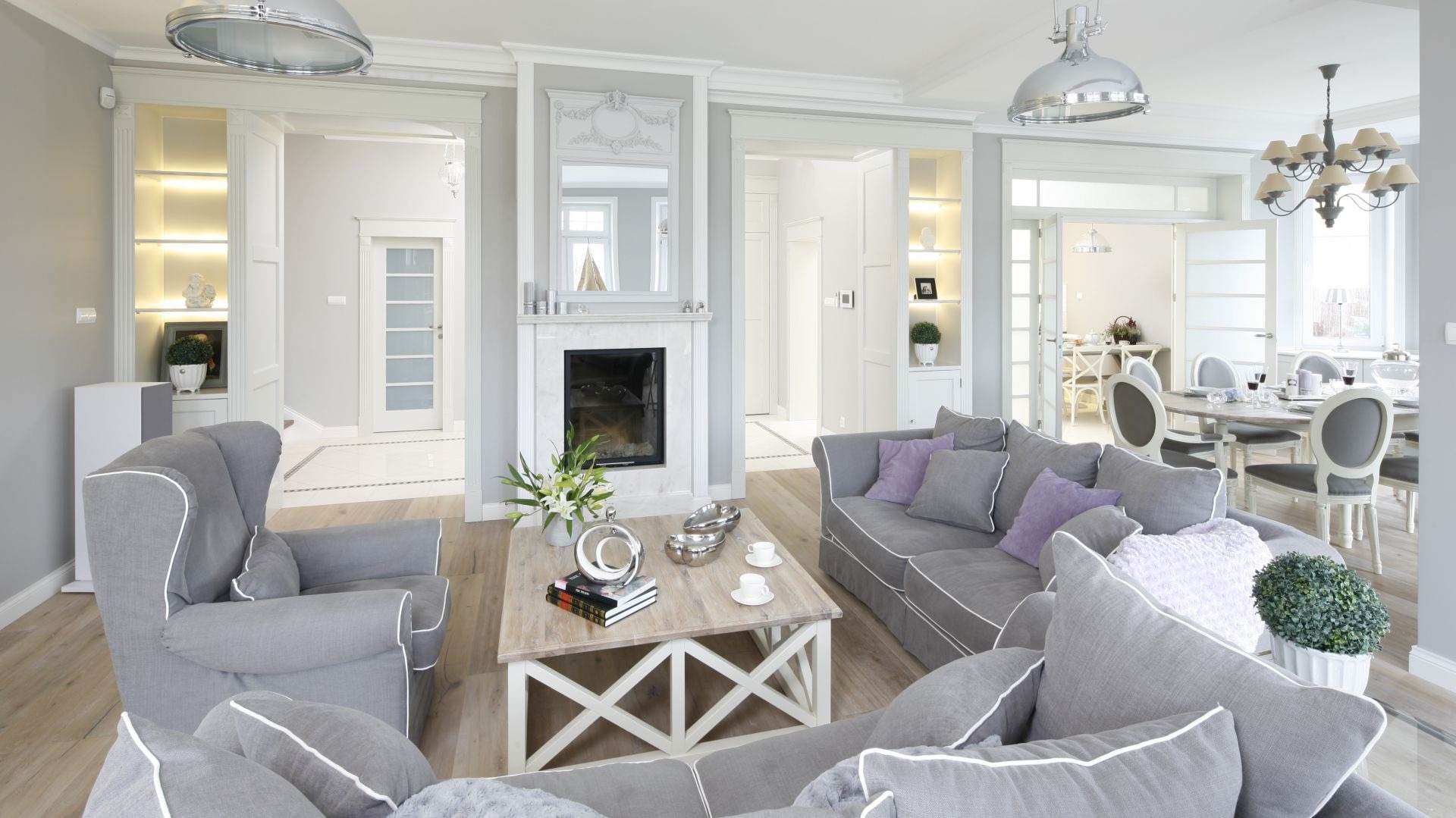 Jasne, przestronne wnętrze salonu z kominkiem, szarymi sofami z fioletowymi poduszkami, drewnianym stolikiem kawowym i widokiem na jadalnię w tle.