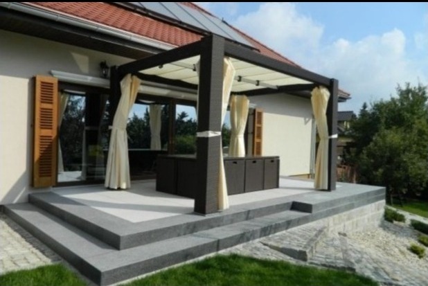Nowoczesna pergola z zasłonami na tarasie z kamiennymi schodami, widok na okno z drewnianymi okiennicami i zadbany ogród.