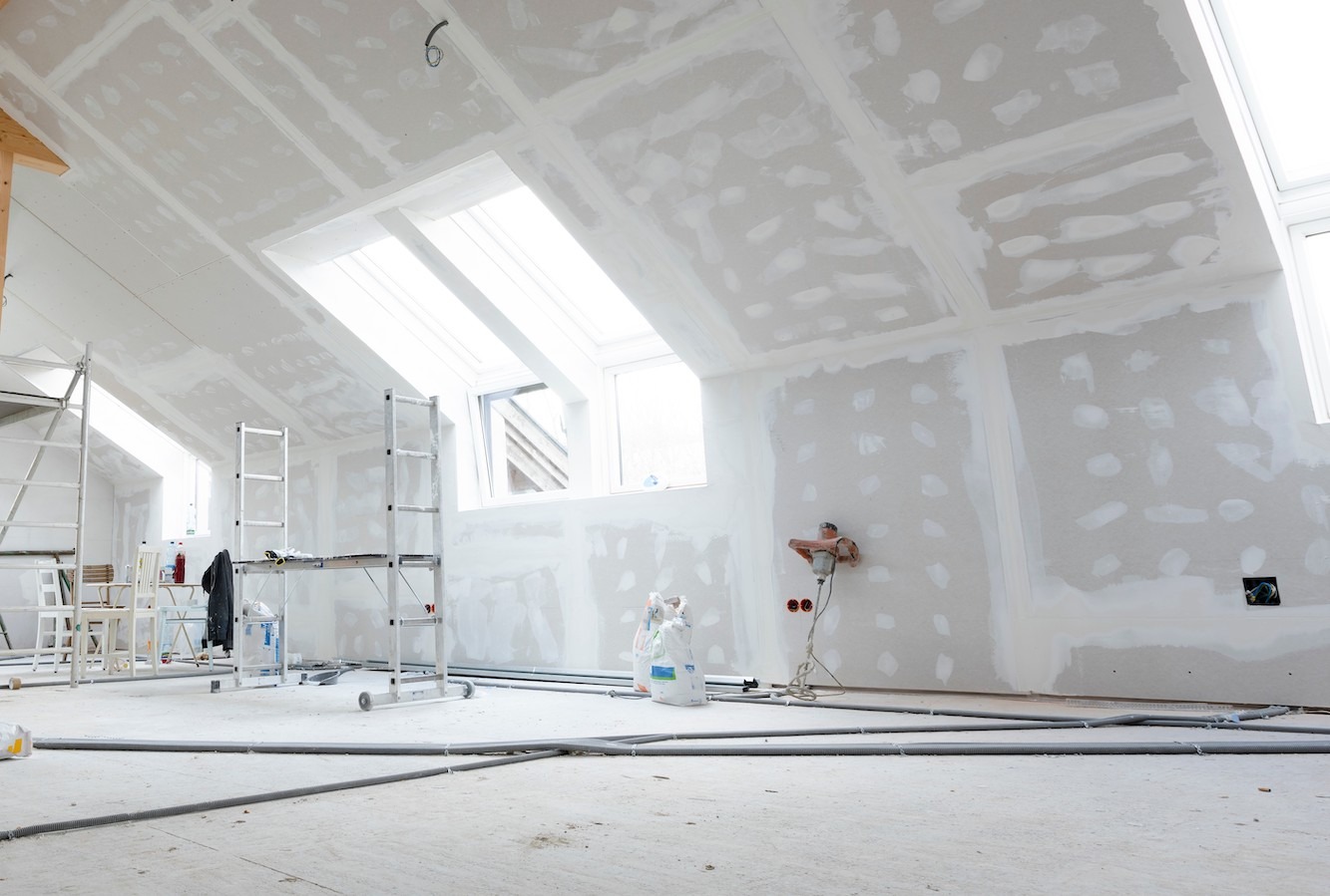 Poddasze w trakcie szpachlowania płyt gipsowo-kartonowych, widoczne rusztowania, worki z gładzią i szlifierka do gipsu na ścianie, okna dachowe w skosach.