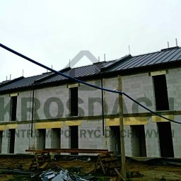 KROSDACH S.C. URSZULA ROSPOND, JAKUB HEJNAR - Szeregówka w trakcie budowy z ciemnym, metalowym pokryciem dachowym i zamontowanymi rynnami, widoczne rusztowania i materiały budowlane.