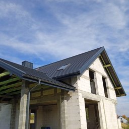 KROSDACH S.C. URSZULA ROSPOND, JAKUB HEJNAR - Niedokończony dom z nowym, ciemnoszarym dachem z blachodachówki, widoczne elementy konstrukcji drewnianej, rynny i okno dachowe, na tle błękitnego nieba z chmurami.