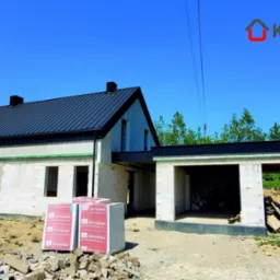 Dom w stanie surowym zamkniętym z ciemnym dachem z blachy na rąbek stojący, widoczne pakiety bloczków Termobet na pierwszym planie.