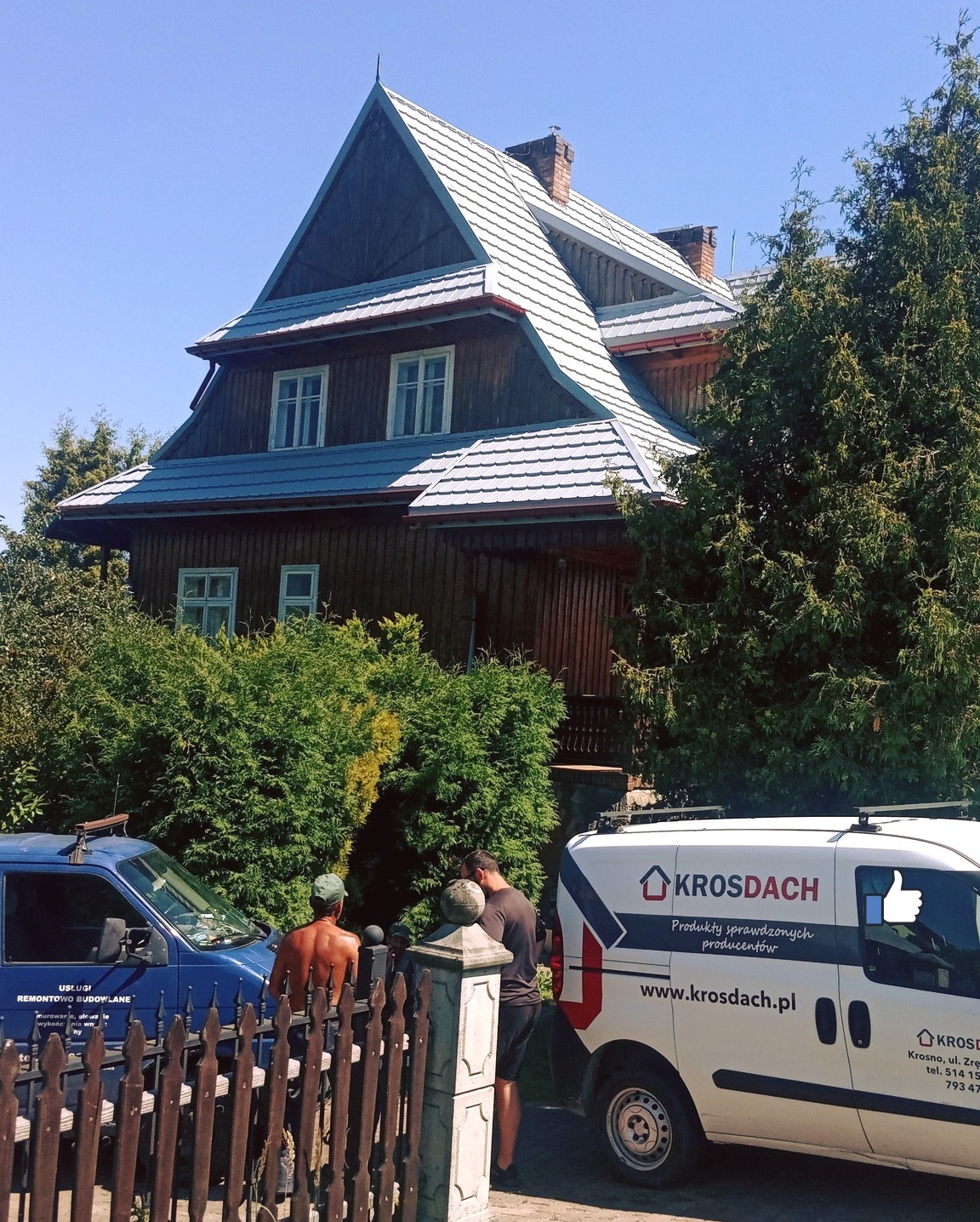 Drewniany dom z dwuspadowym dachem pokrytym szarą dachówką karpiówką, widoczny samochód firmowy z logo na tle zieleni i dwóch mężczyzn za ogrodzeniem.