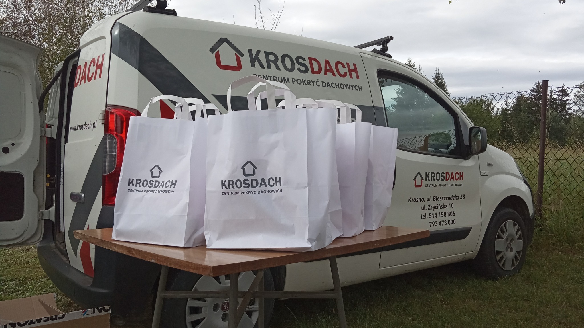 Reklamowy samochód firmy z Krosna, specjalizującej się w pokryciach dachowych, zaparkowany na zewnątrz. Na stole obok niego leżą białe torby z logo firmy.