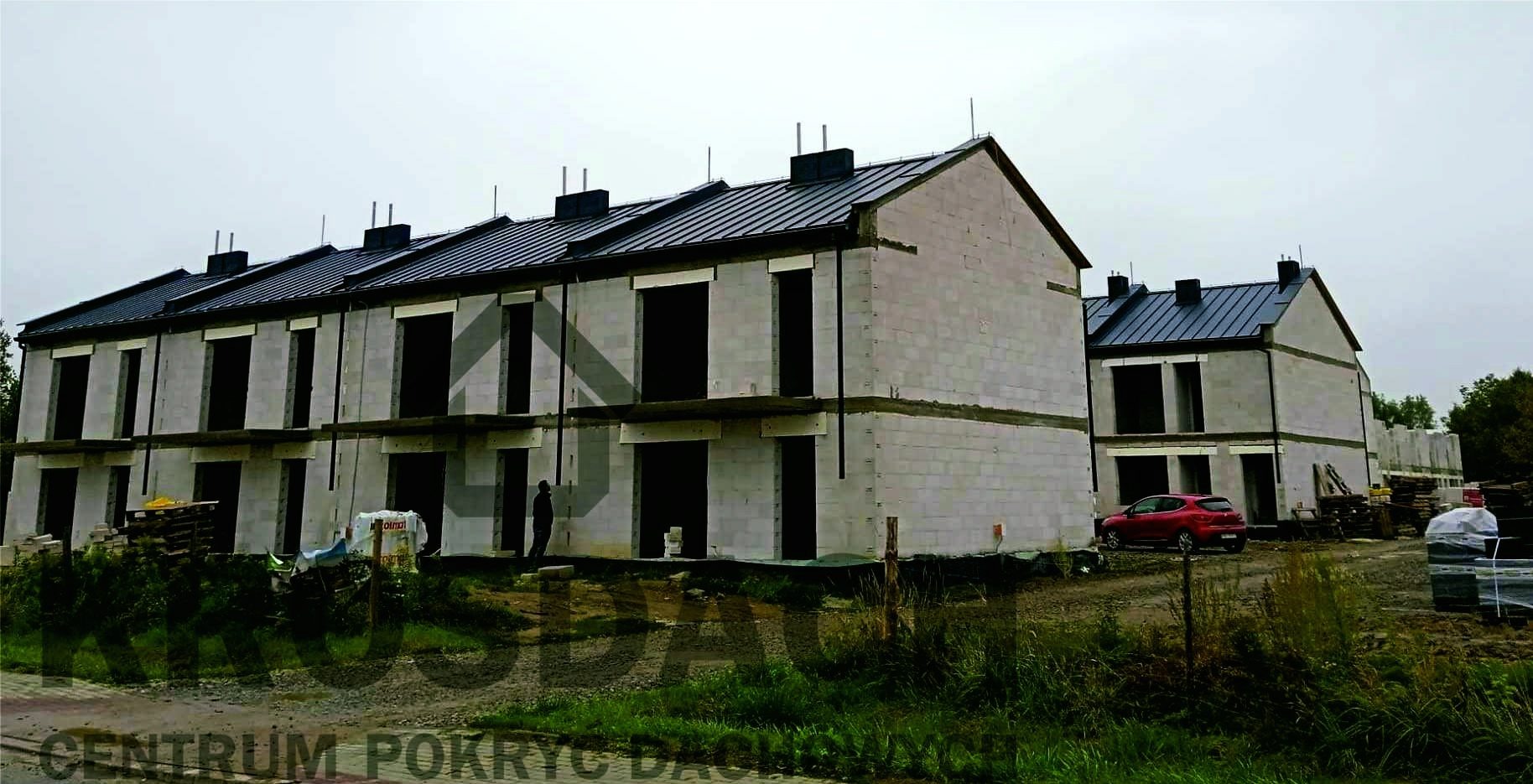 Szeregówka w trakcie budowy z ukończonym dachem z blachy na rąbek stojący w kolorze grafitowym, widoczne kominy i orynnowanie, stan surowy zamknięty.
