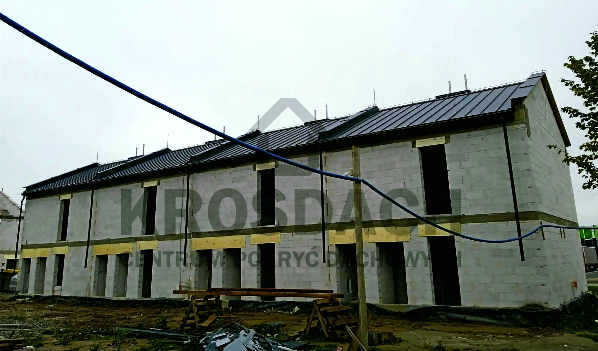 Szeregówka w trakcie budowy z ciemnym, metalowym pokryciem dachowym i zamontowanymi rynnami, widoczne rusztowania i materiały budowlane.
