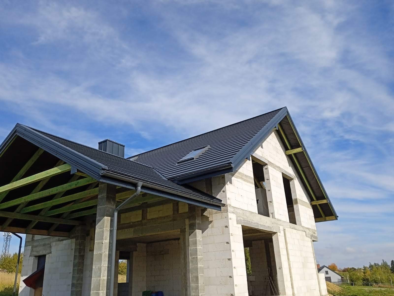 Niedokończony dom z nowym, ciemnoszarym dachem z blachodachówki, widoczne elementy konstrukcji drewnianej, rynny i okno dachowe, na tle błękitnego nieba z chmurami.