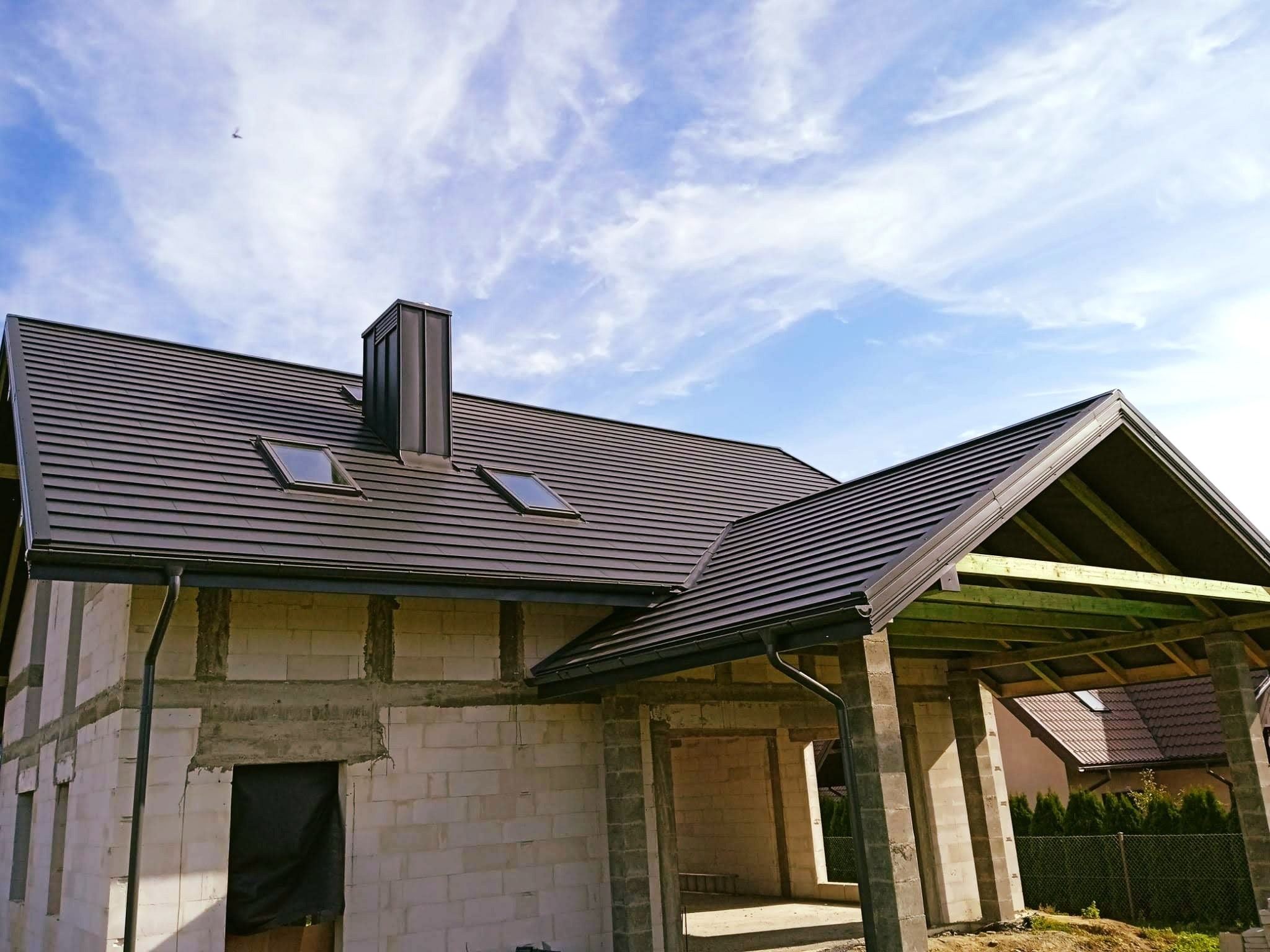 Dom w trakcie budowy z nowym, ciemnym dachem z dachówkami, rynnami i oknami dachowymi, widoczny komin i fragment konstrukcji werandy z zielonymi belkami, na tle błękitnego nieba z chmurami.