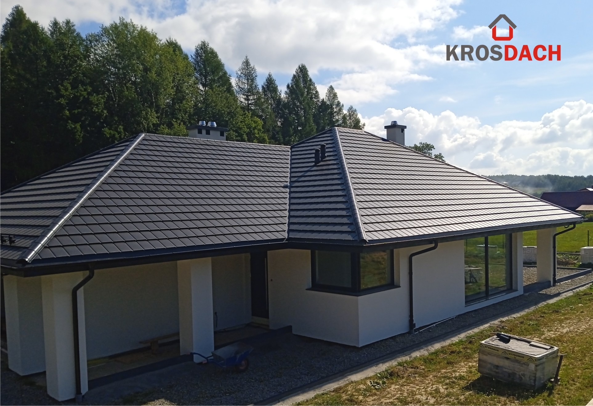 Nowoczesny dom jednorodzinny z ciemnoszarą dachówką modułową, widok z boku, na dachu komin i logo firmy Krosdach