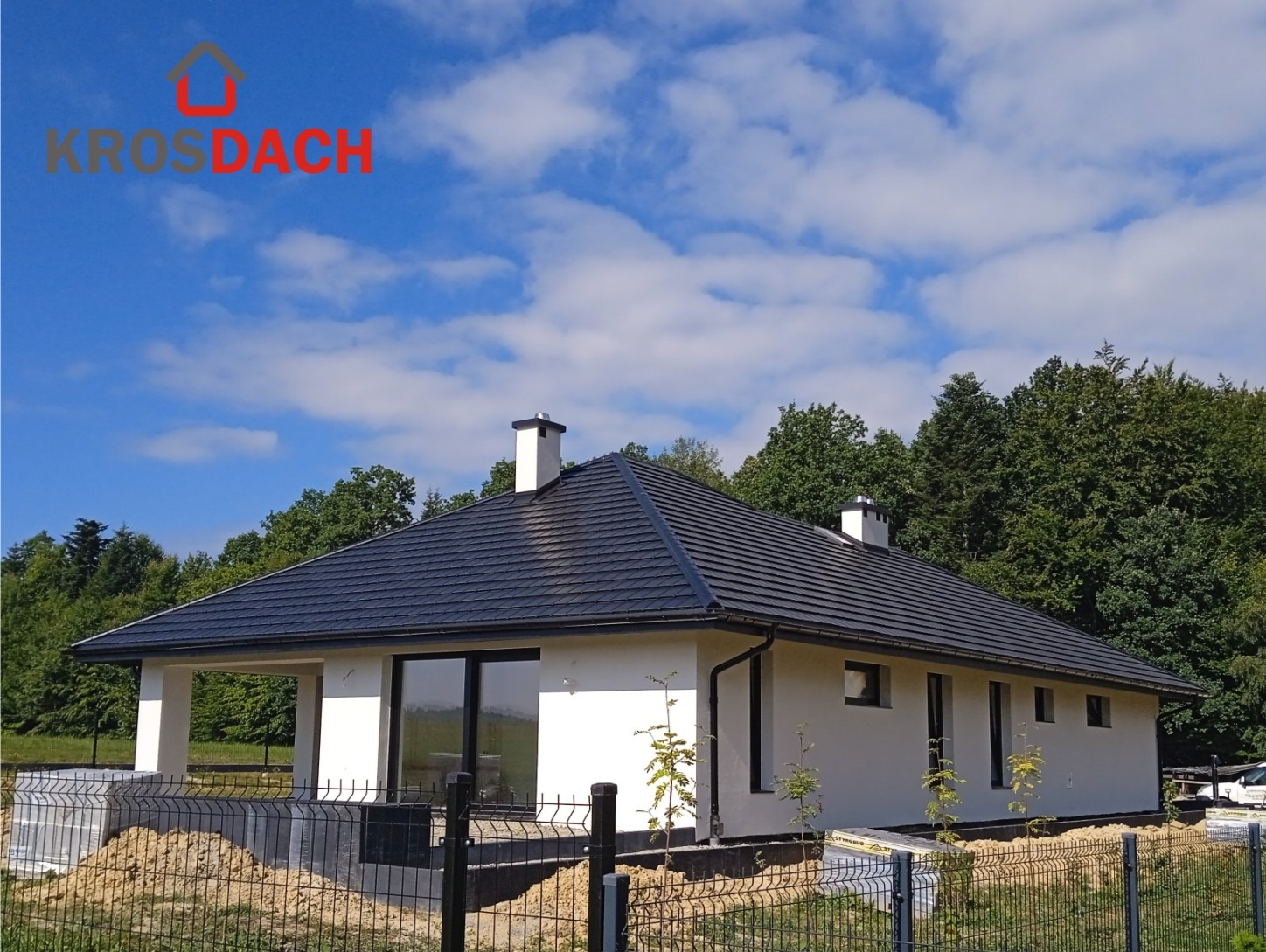 Nowoczesny dom jednorodzinny z ciemną, płaską dachówką, białą elewacją i czarnymi ramami okiennymi, widok zza ogrodzenia w słoneczny dzień.